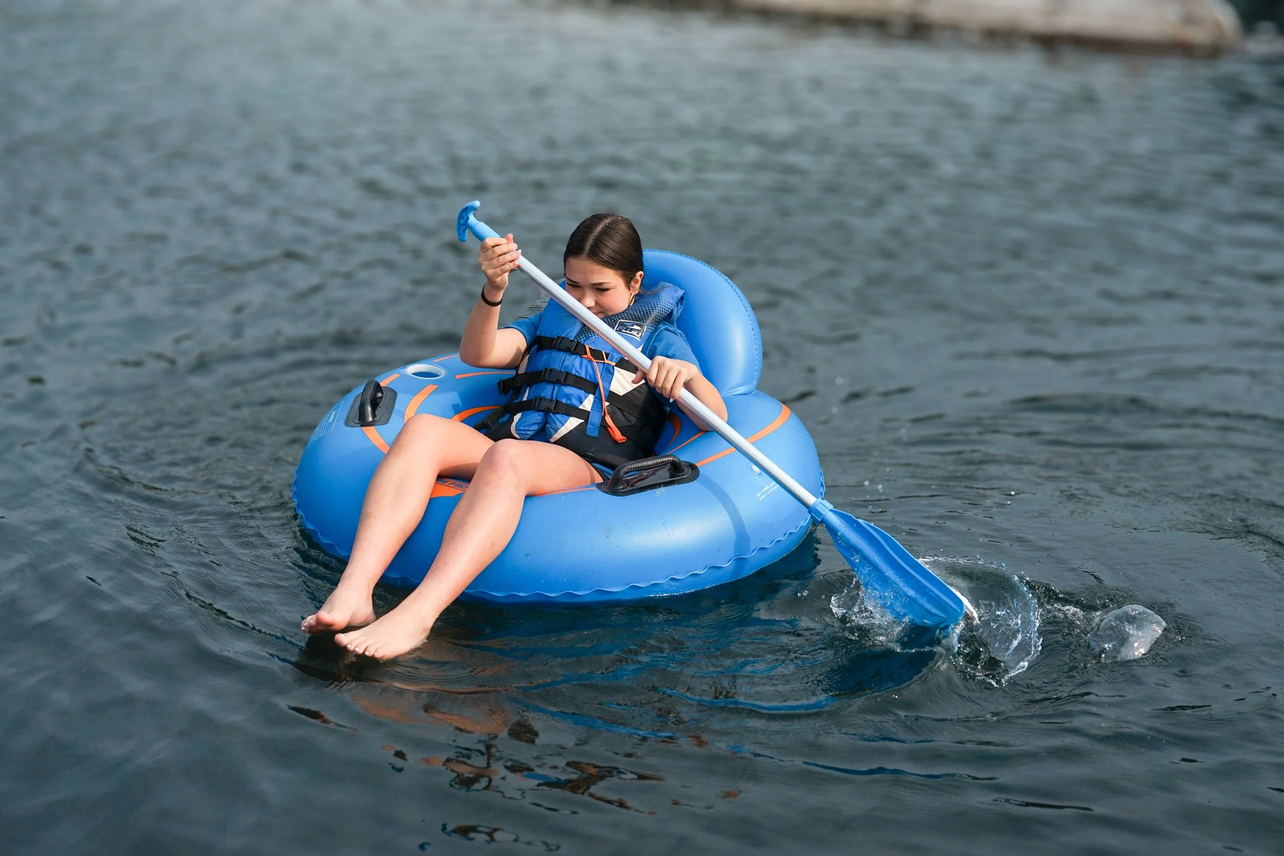 A young girl smiling and relaxing on a blue inflatable tube in the river, wearing a life jacket with her legs in the water.