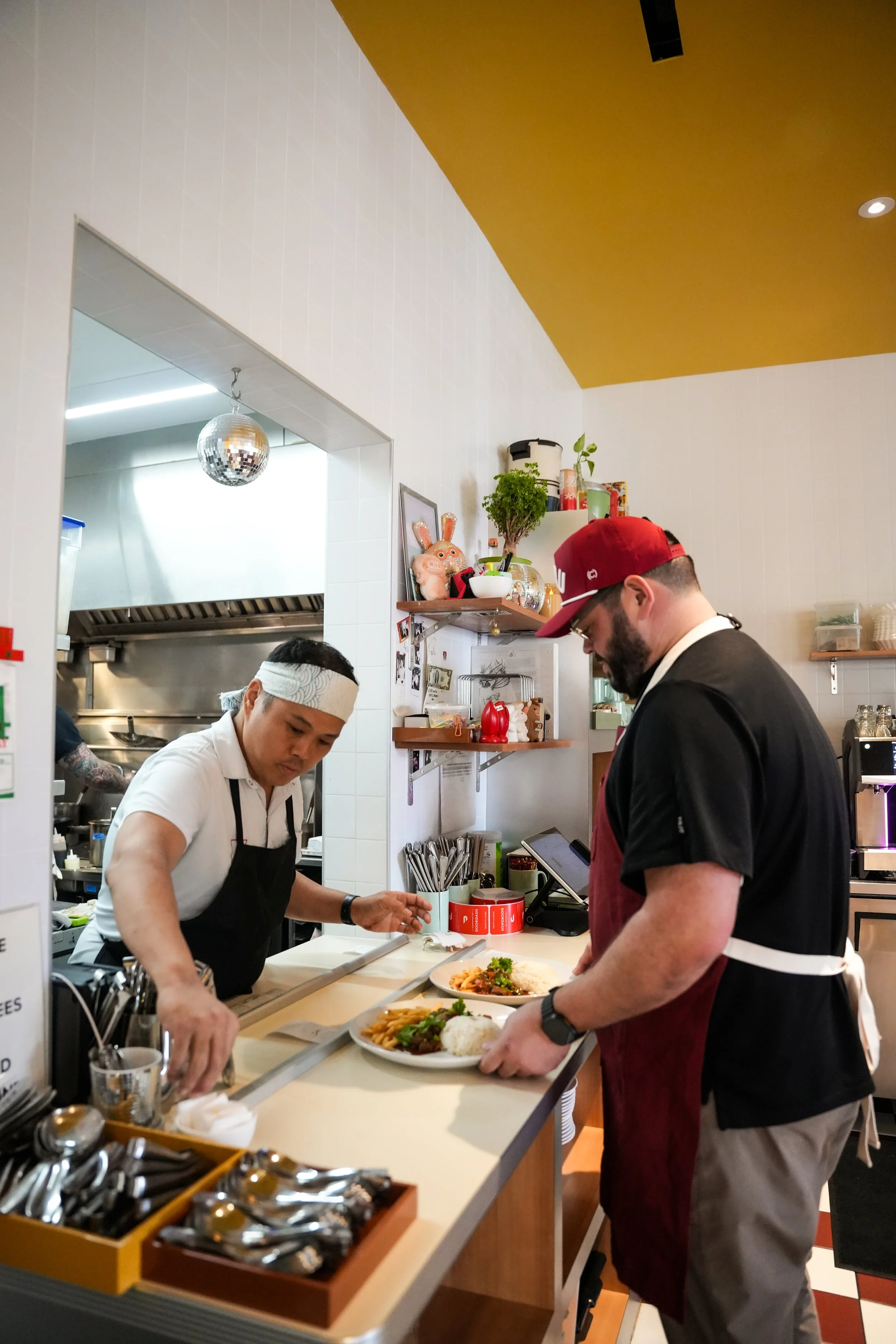 Two male chefs in a restaurant kitchen preparing dishes while a customer places an order at the counter. The kitchen features shelves with plants, decor, and kitchen tools, and a disco ball hanging from the ceiling.