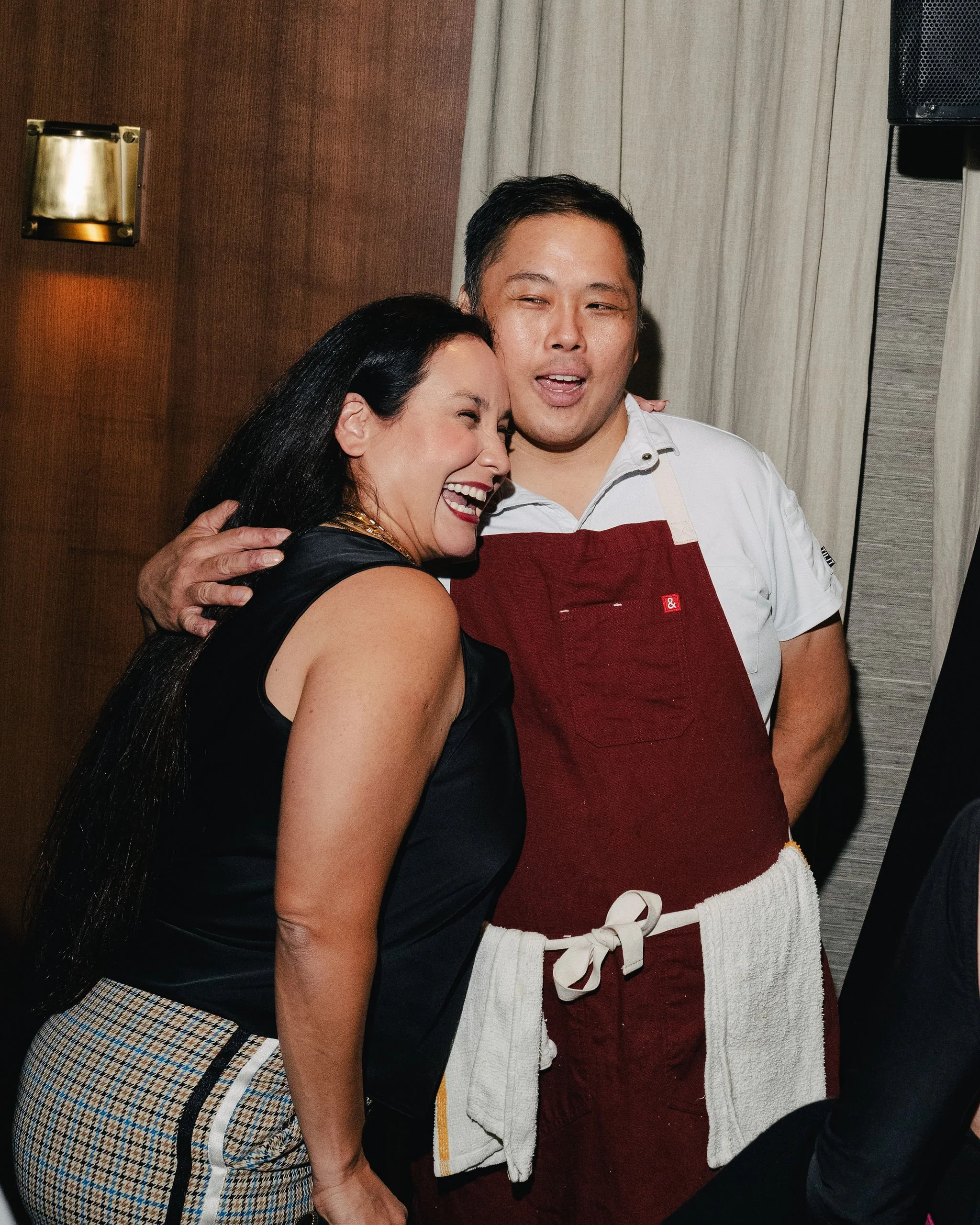 Two women and a man at a social gathering. The woman with long dark hair is smiling and hugging the man, who is wearing a white shirt and a red apron. The woman is wearing a sleeveless black top and plaid skirt. The man is in front of beige curtains, with a wall sconce and part of a speaker visible in the background.