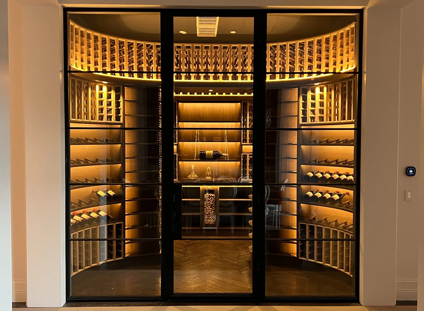 A modern wine cellar with glass doors, warm wooden shelving, and integrated lighting for wine storage and display.