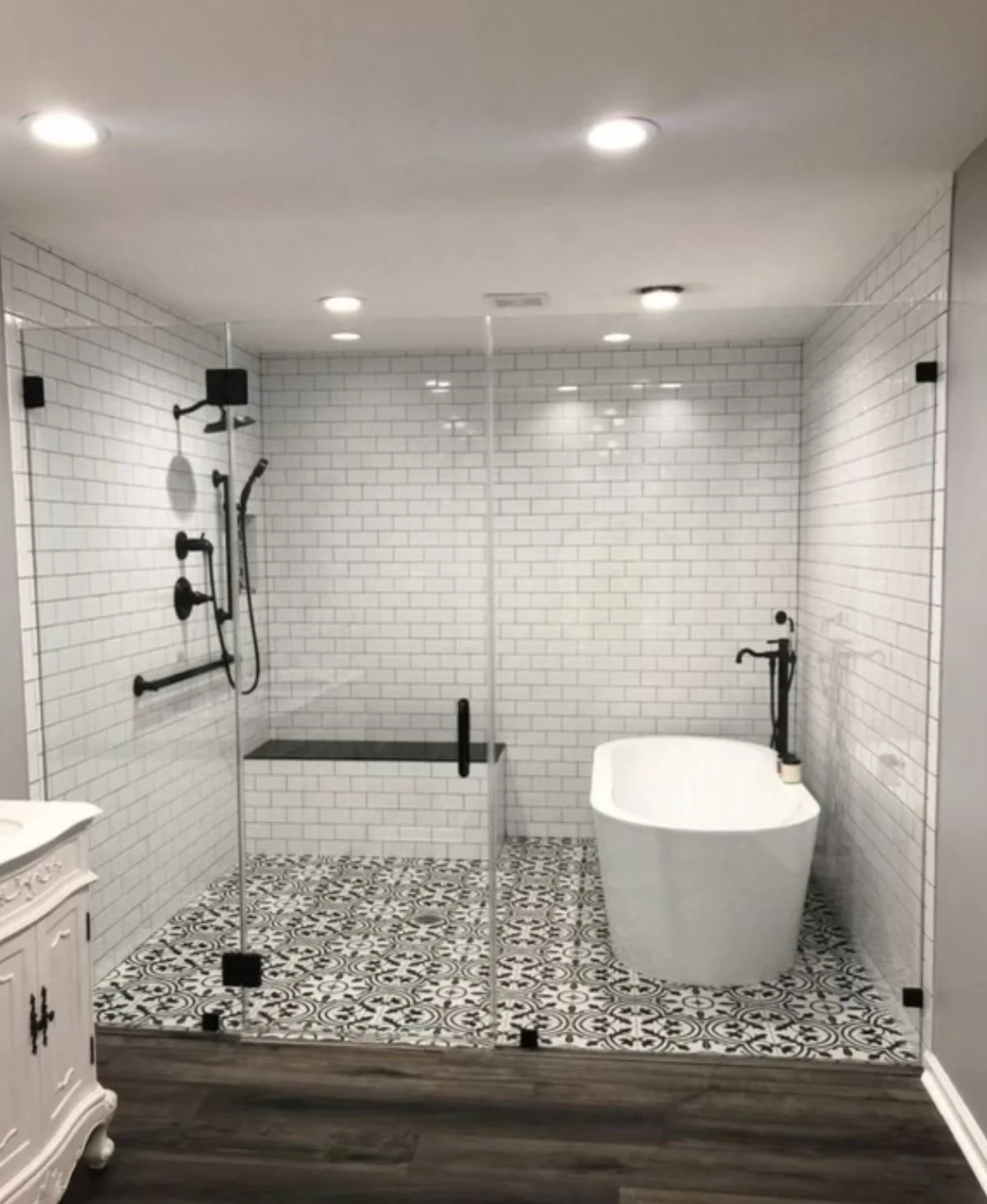 Modern walk-in shower with glass enclosure, black fixtures, white subway tile walls, patterned floor tiles, and a white freestanding bathtub.