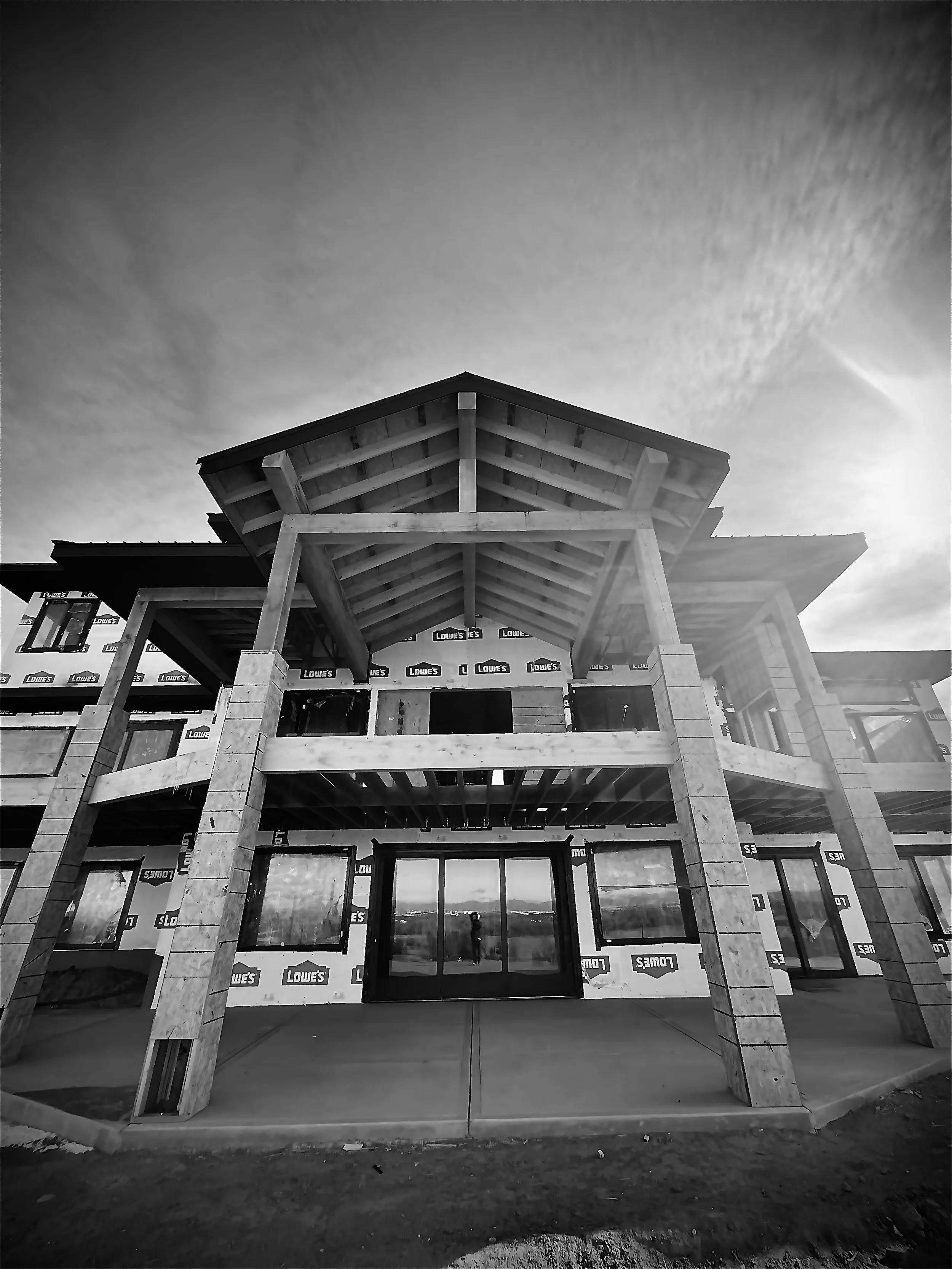 Under-construction building with wooden framework and concrete columns, reflecting in glass doors, with cloudy sky overhead.