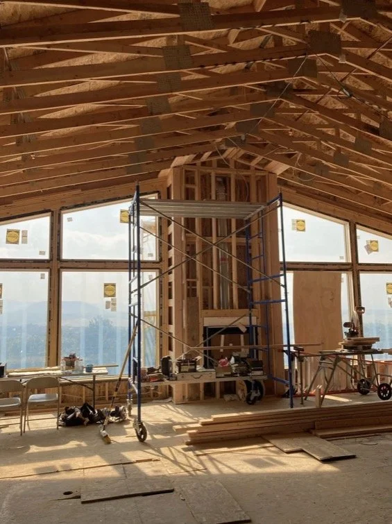 Interior of a building under construction with exposed wooden framing, scaffolding, and large windows showing an outdoor view.