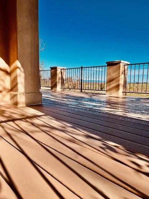 View from a wooden deck with shadows cast by a nearby structure, overlooking a landscape with a railing and a clear blue sky.