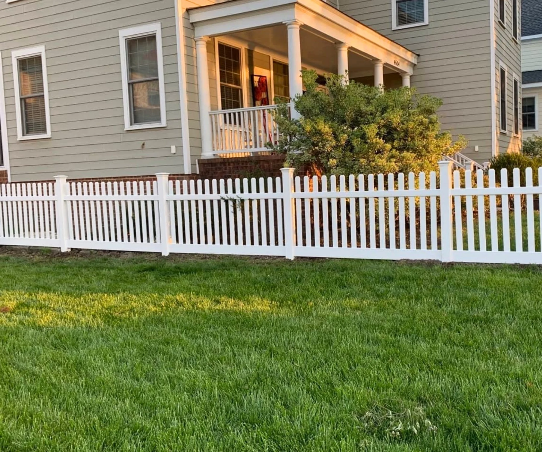 Exterior view of a two-story house with beige siding, white window frames, and a front porch with white columns, a white vinyl picket fence, and green lawn in the foreground.