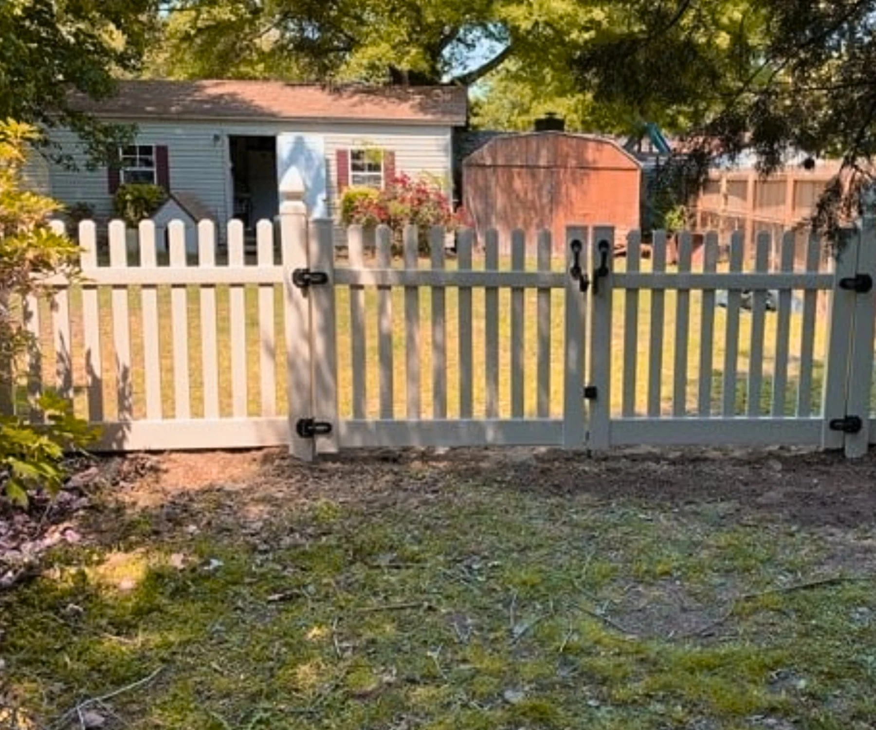 White picket fence with gate, wooden shed, small house with flower porch, trees in yard, sunny weather