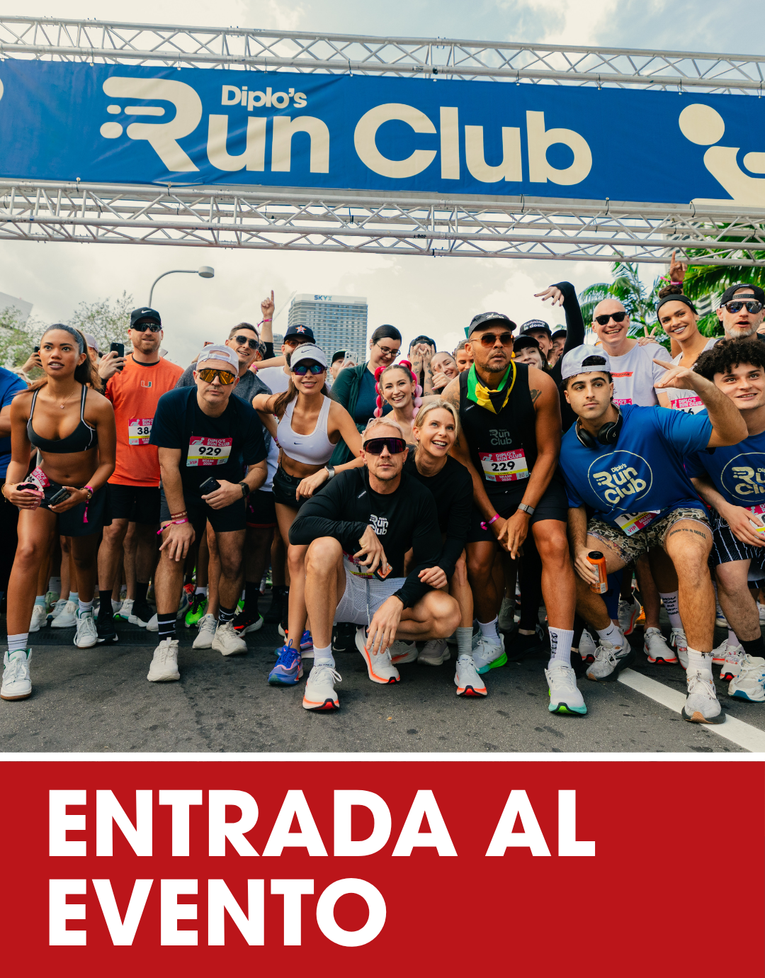 Large crowd gathered at an outdoor event with a stage featuring colorful banners, flags, and graphic icons of running, cycling, and swimming, with performers and speakers on stage and tall buildings in the background. The bottom of the image has an orange banner with white text reading 'EVENT ENTRY'.