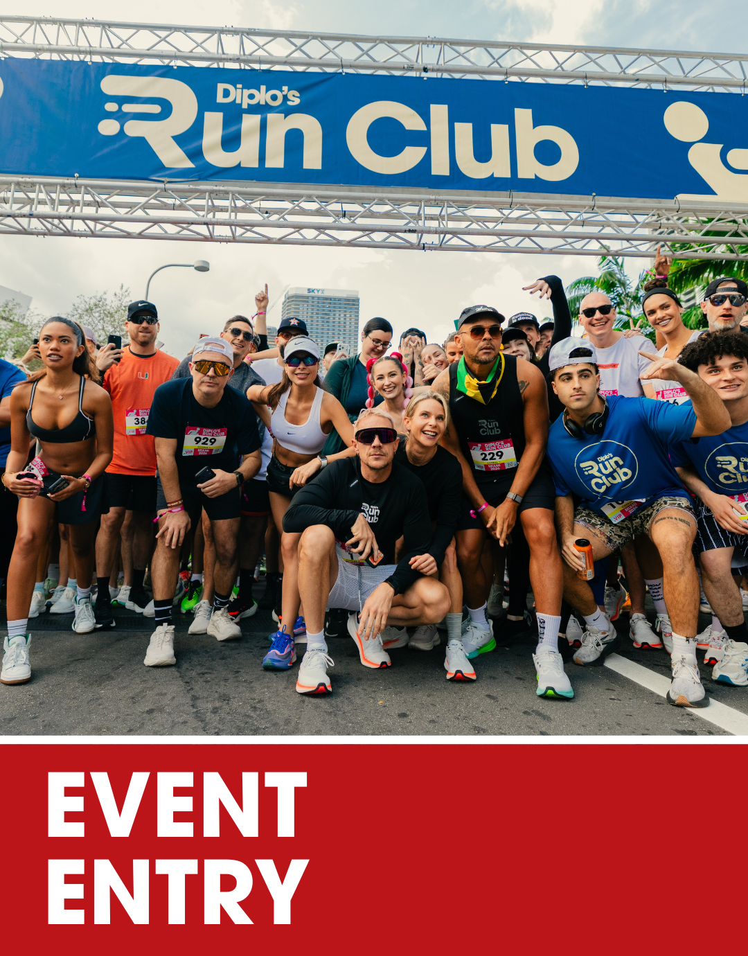 Large crowd gathered at an outdoor event with a stage featuring colorful banners, flags, and graphic icons of running, cycling, and swimming, with performers and speakers on stage and tall buildings in the background. The bottom of the image has an orange banner with white text reading 'EVENT ENTRY'.