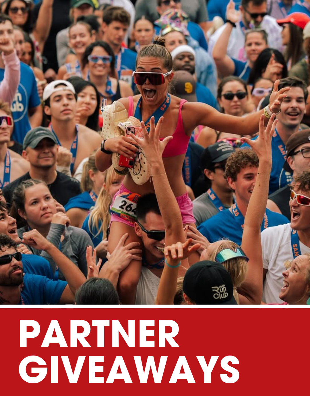 A man wearing sunglasses and a black long-sleeve shirt is at an outdoor event, holding a medal and surrounded by boxes of supplements labeled 'Golden Mind' and 'Symbiotika'. There are baskets filled with more supplement packets on the table, with a medium-sized crowd and city buildings in the background. The text 'Partner Giveaways' is displayed in bold white letters on an orange banner at the bottom.