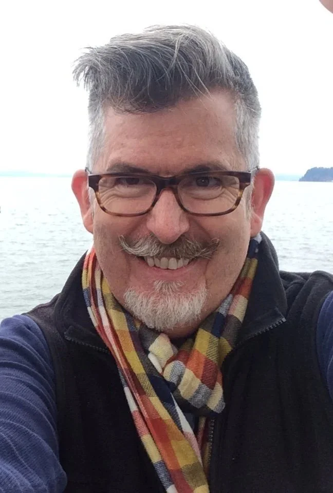 Photo of Tom Cotter smiling, wearing a colorful scarf and a black vest, outdoors near water with an island in the background.