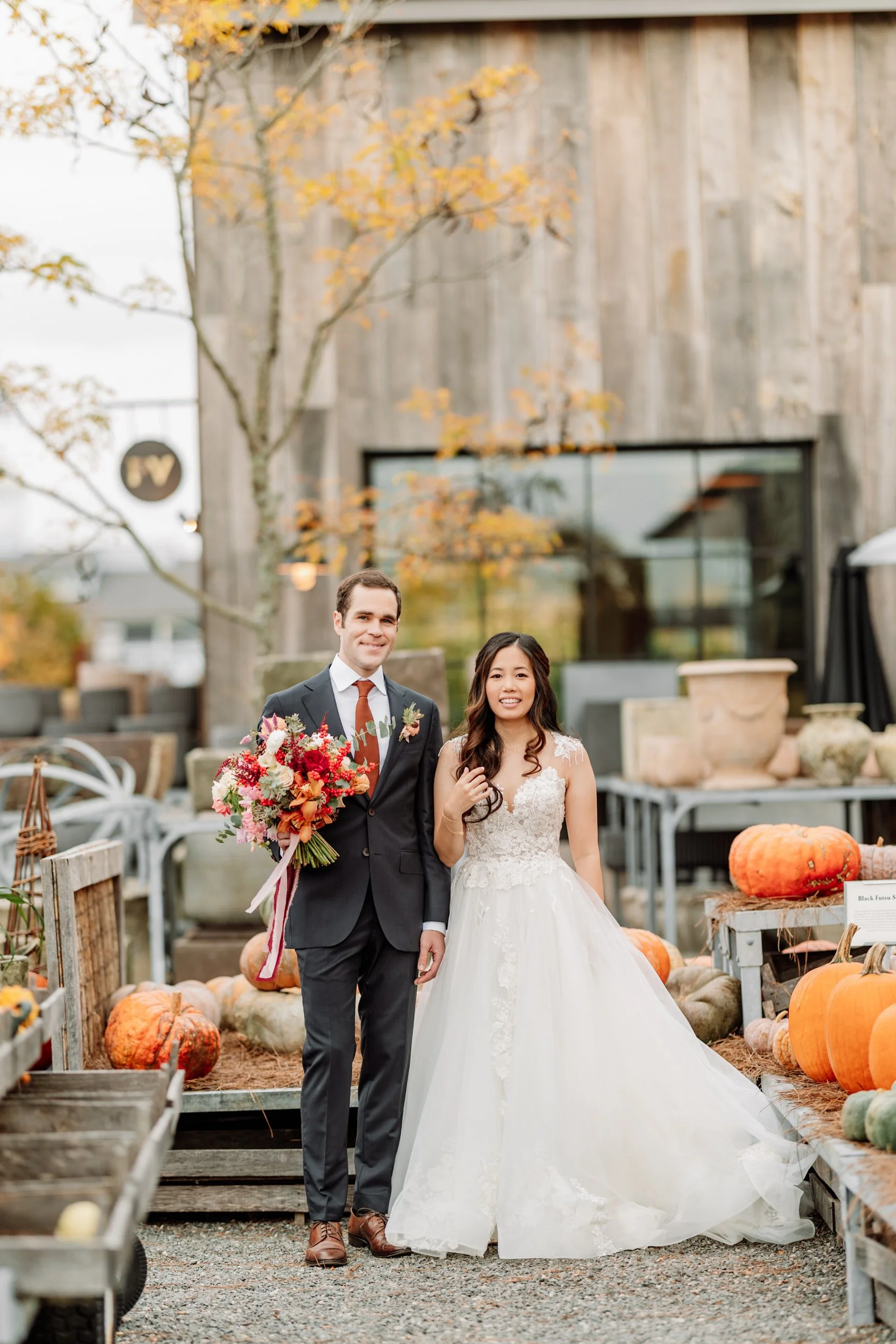 A newlywed couple posing at a pumpkin patch. The groom is in a dark suit with a red tie and boutonniere, holding a colorful bouquet. The bride is in a white lace wedding gown with a full skirt. Autumn leaves and pumpkins decorate the scene.