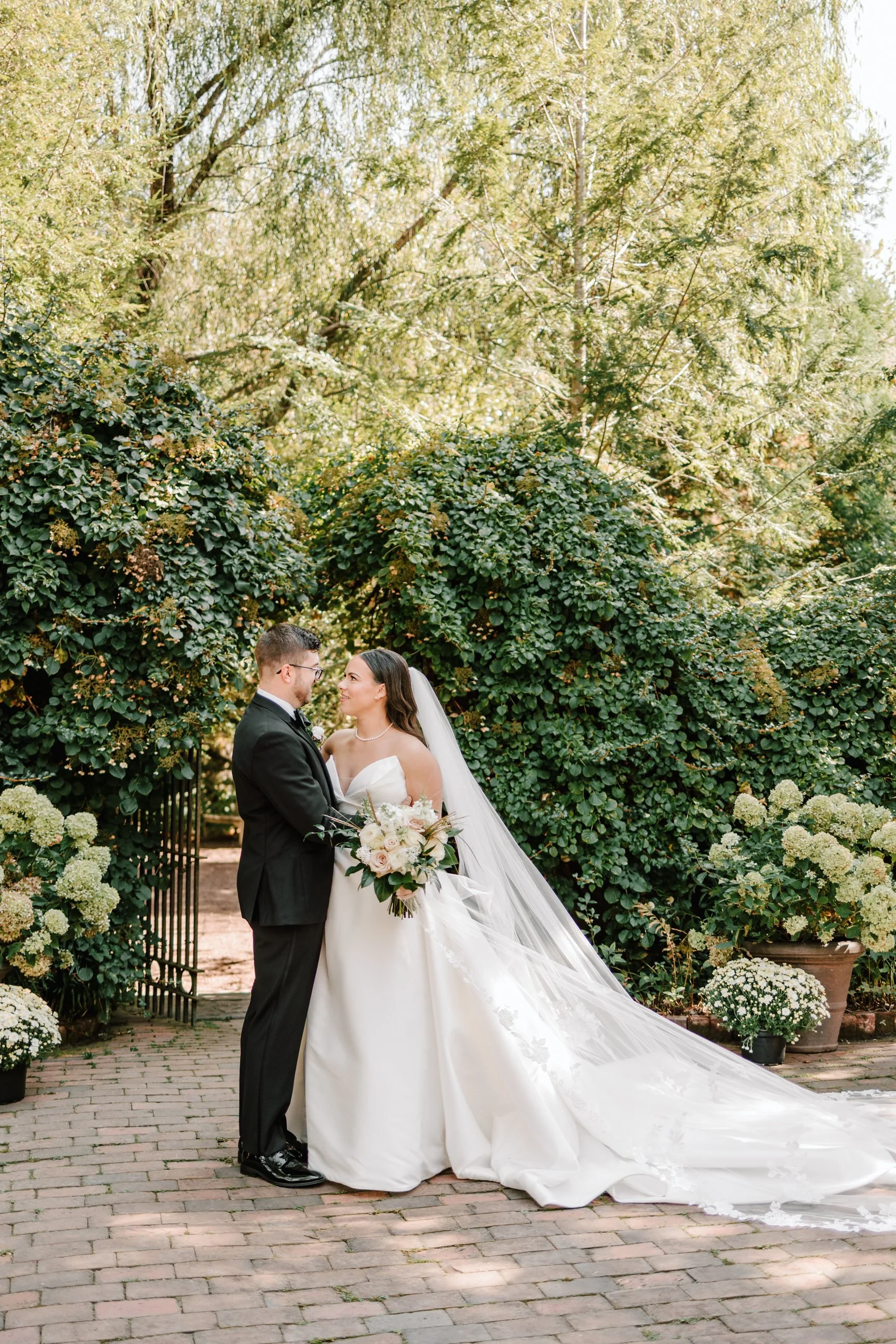 A bride and groom standing close together outdoors, holding a bouquet, surrounded by greenery and blooming flowers.