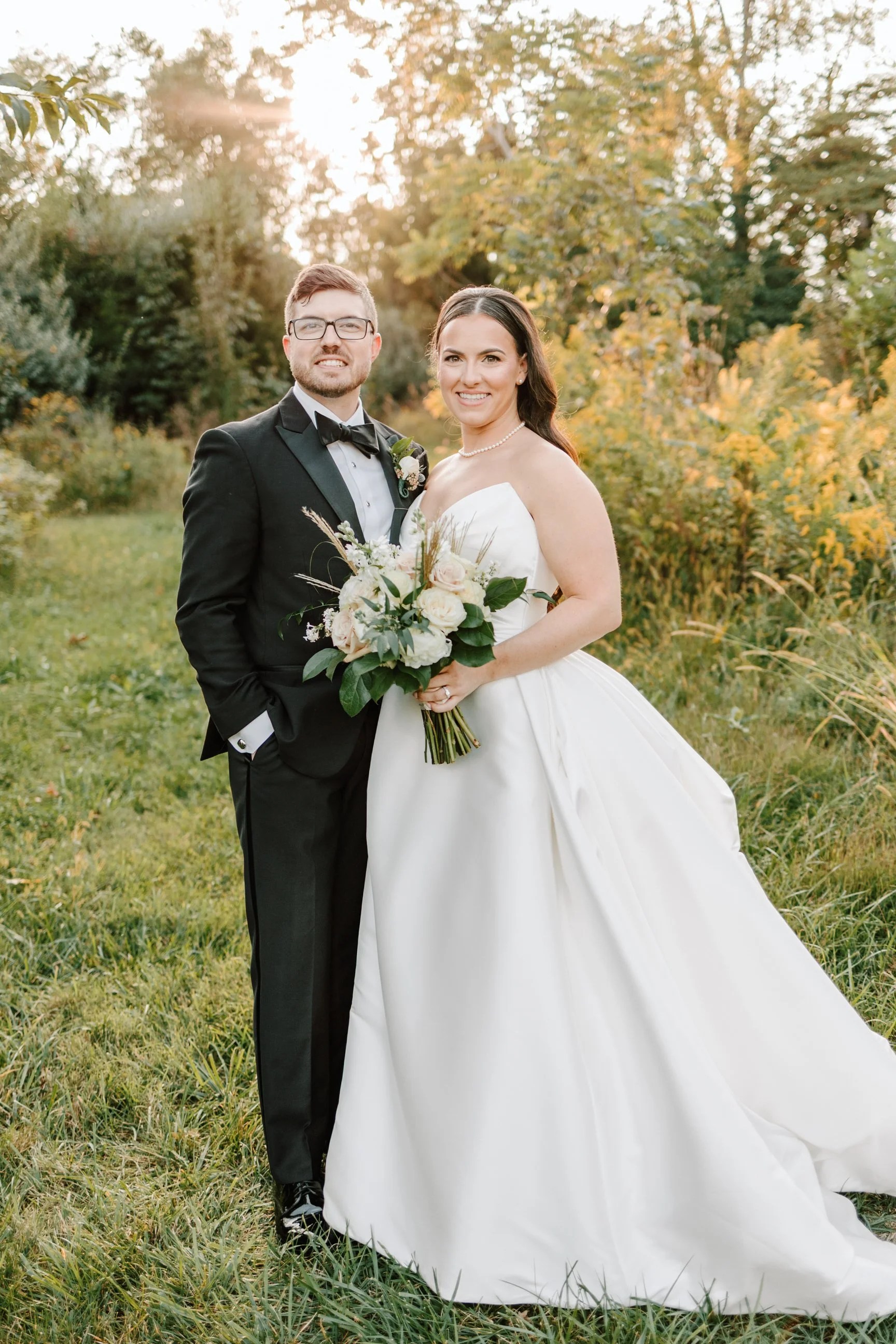 Bride and groom standing outdoors on a grassy field with trees in the background, sunset lighting, dressed in wedding attire, smiling at the camera, the bride holding a bouquet of flowers.