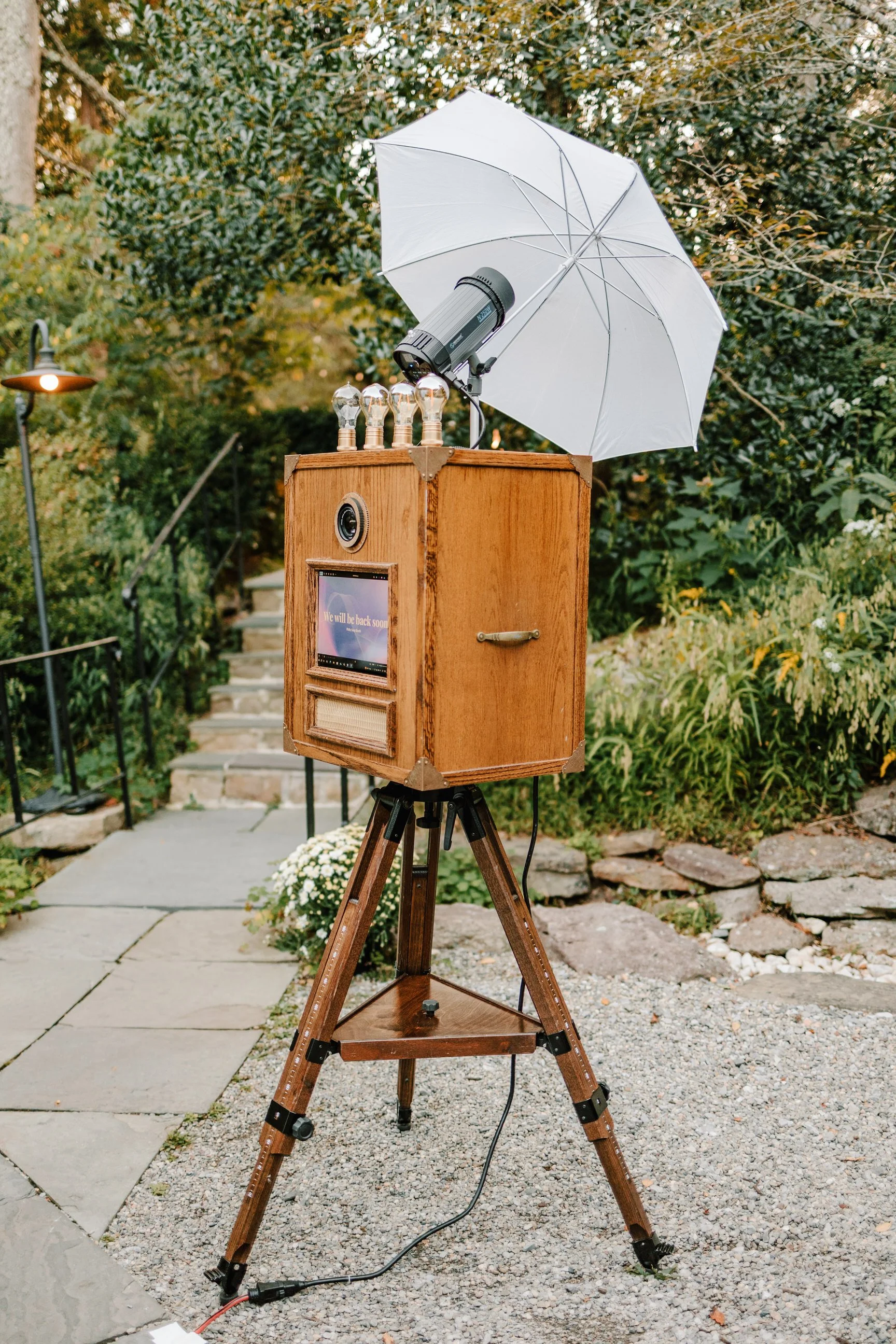 A photo of a vintage photo booth setup outdoors with a wooden box, a white umbrella, and a camera. The booth has a display screen that shows "We will be back soon", and there are four light bulbs on top of the box. The scene is set in a garden with g