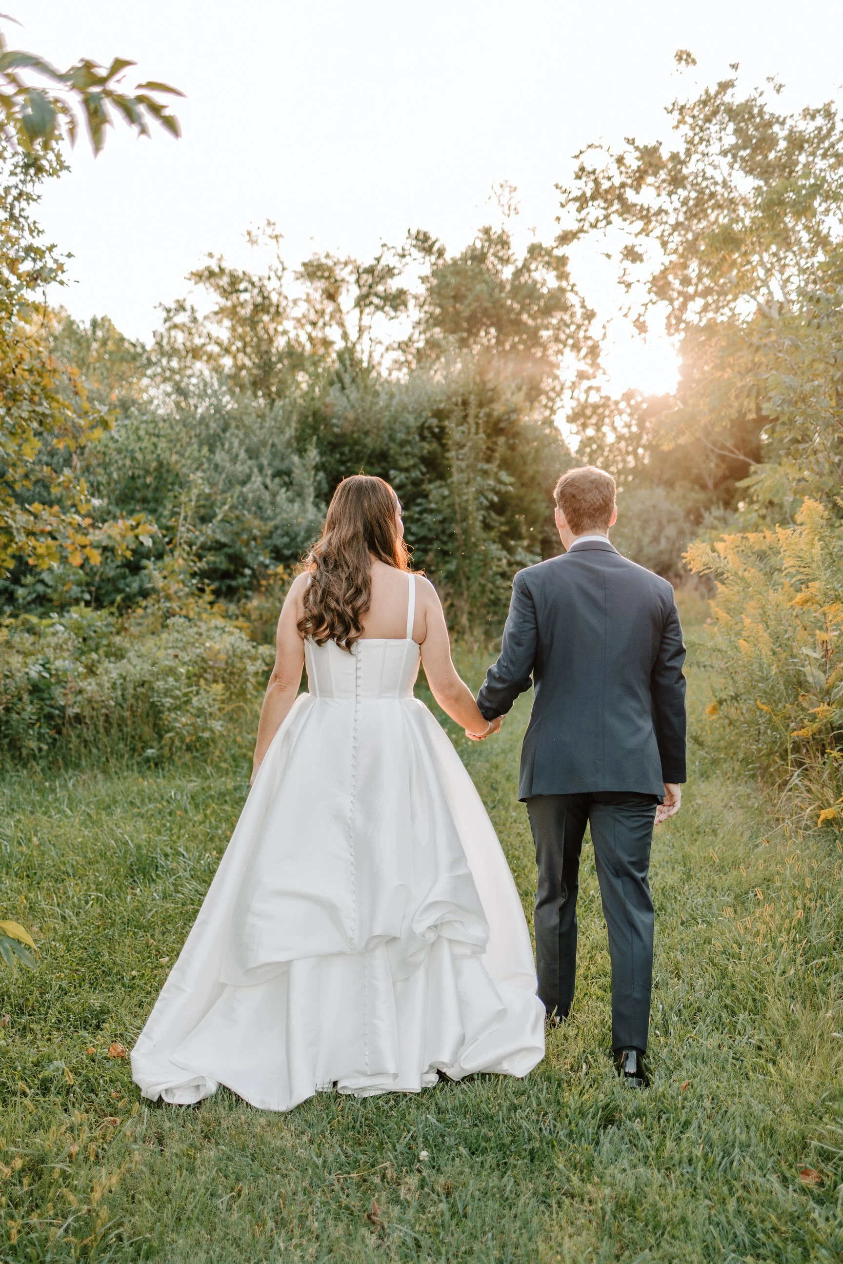 A bride and groom holding hands, walking on a grassy path through a wooded area at sunset, with the sunlight filtering through the trees.
