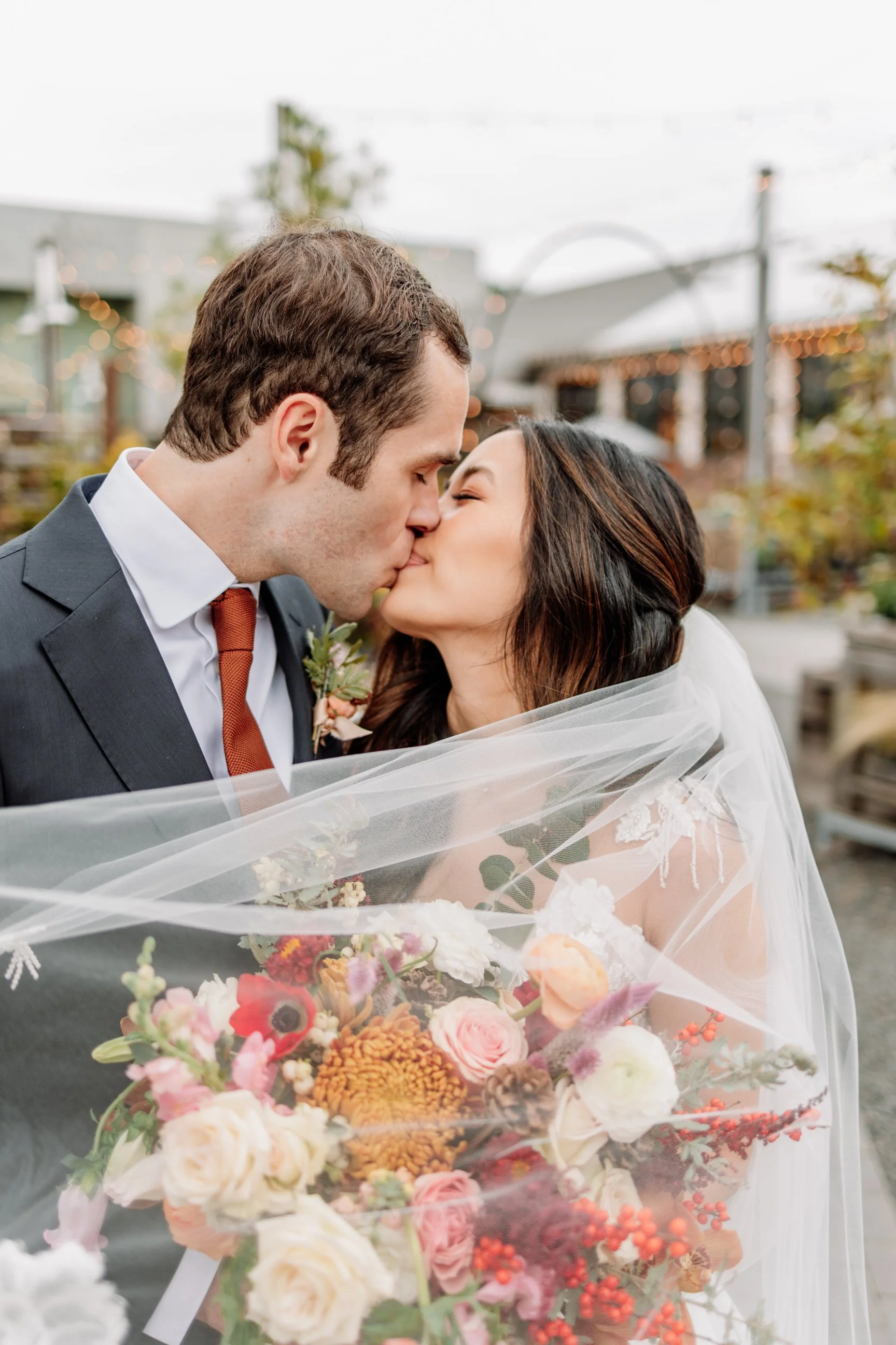 A newlywed couple sharing a kiss outdoors, with the bride holding a bouquet of colorful flowers and the groom in a dark suit with a rust-colored tie.