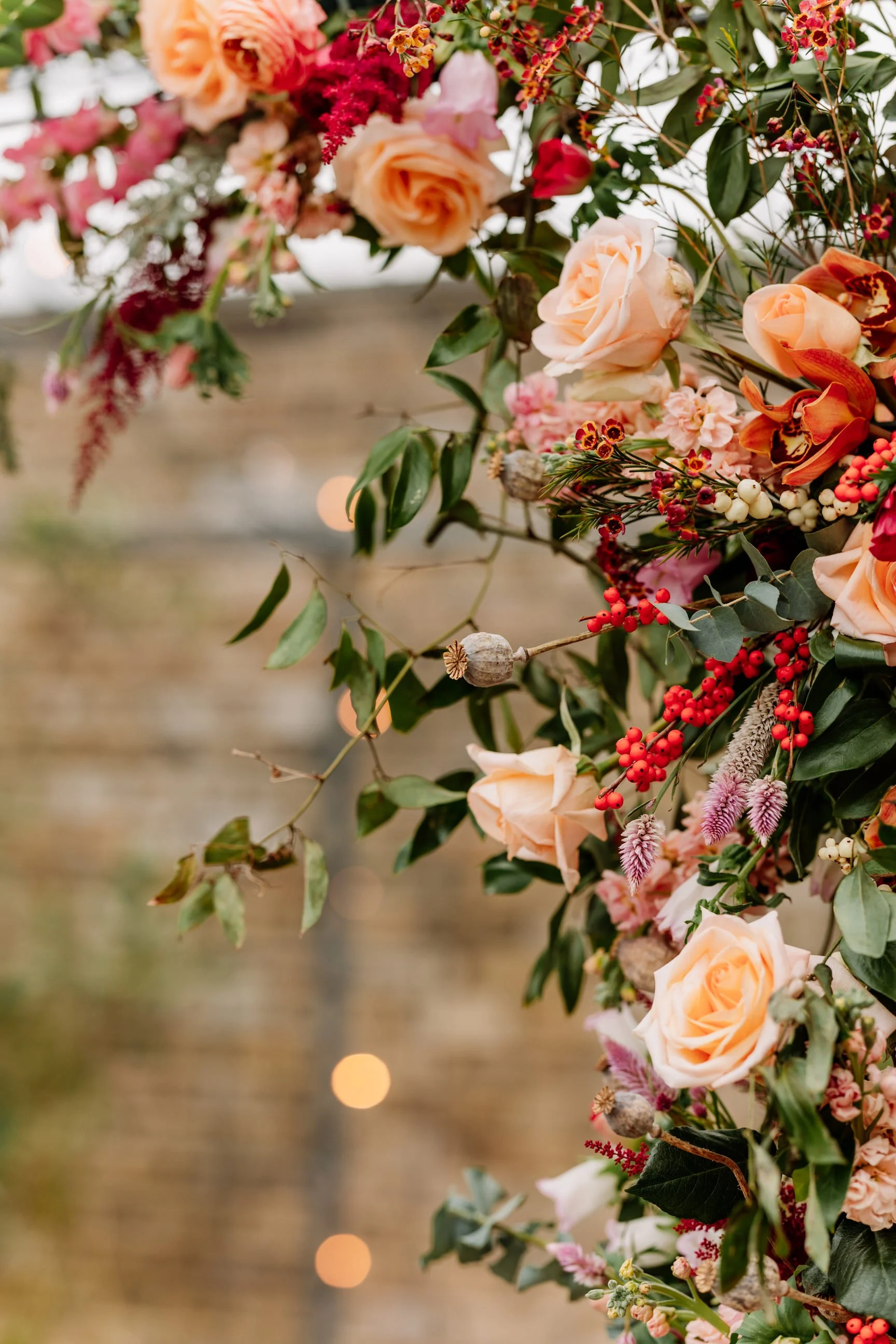 A hanging floral arrangement featuring pink roses, red berries, small pink flowers, and green leaves, with blurred lights in the background.