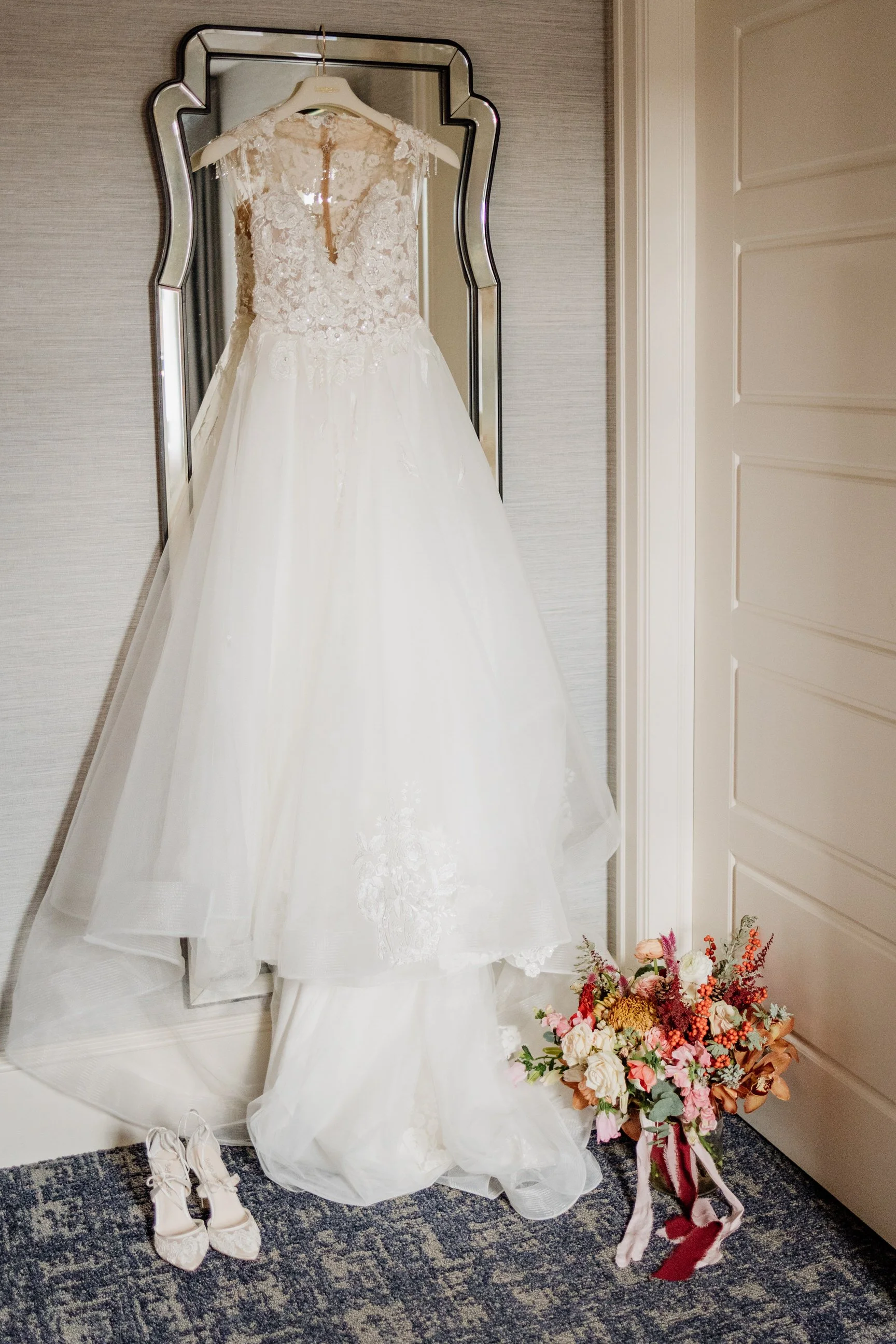 Wedding dress hanging on a mirror, a pair of shoes on the floor, and a bouquet of flowers on the carpet.