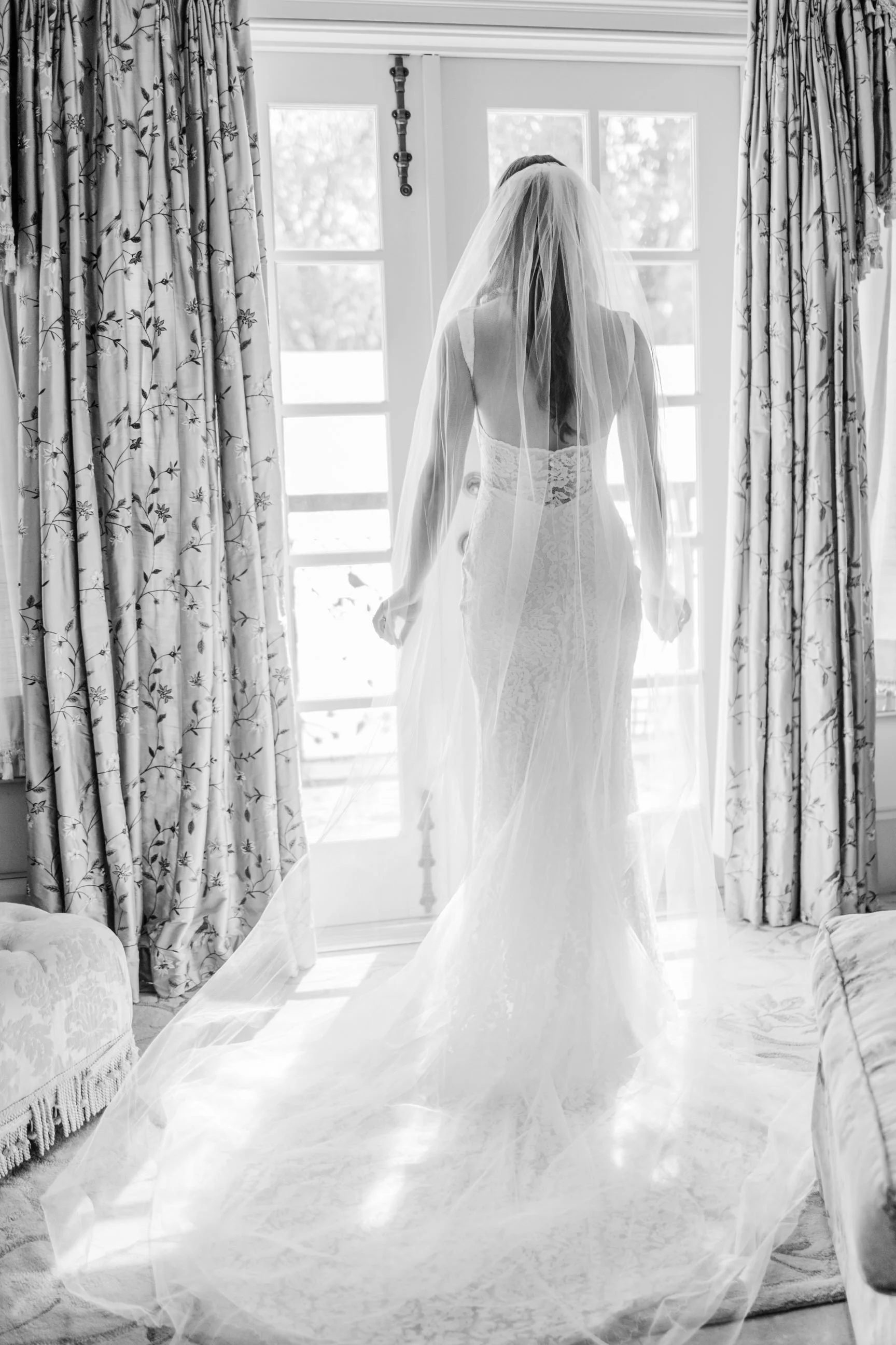 A bride in a wedding dress and veil standing by a window, back view, black and white photo.