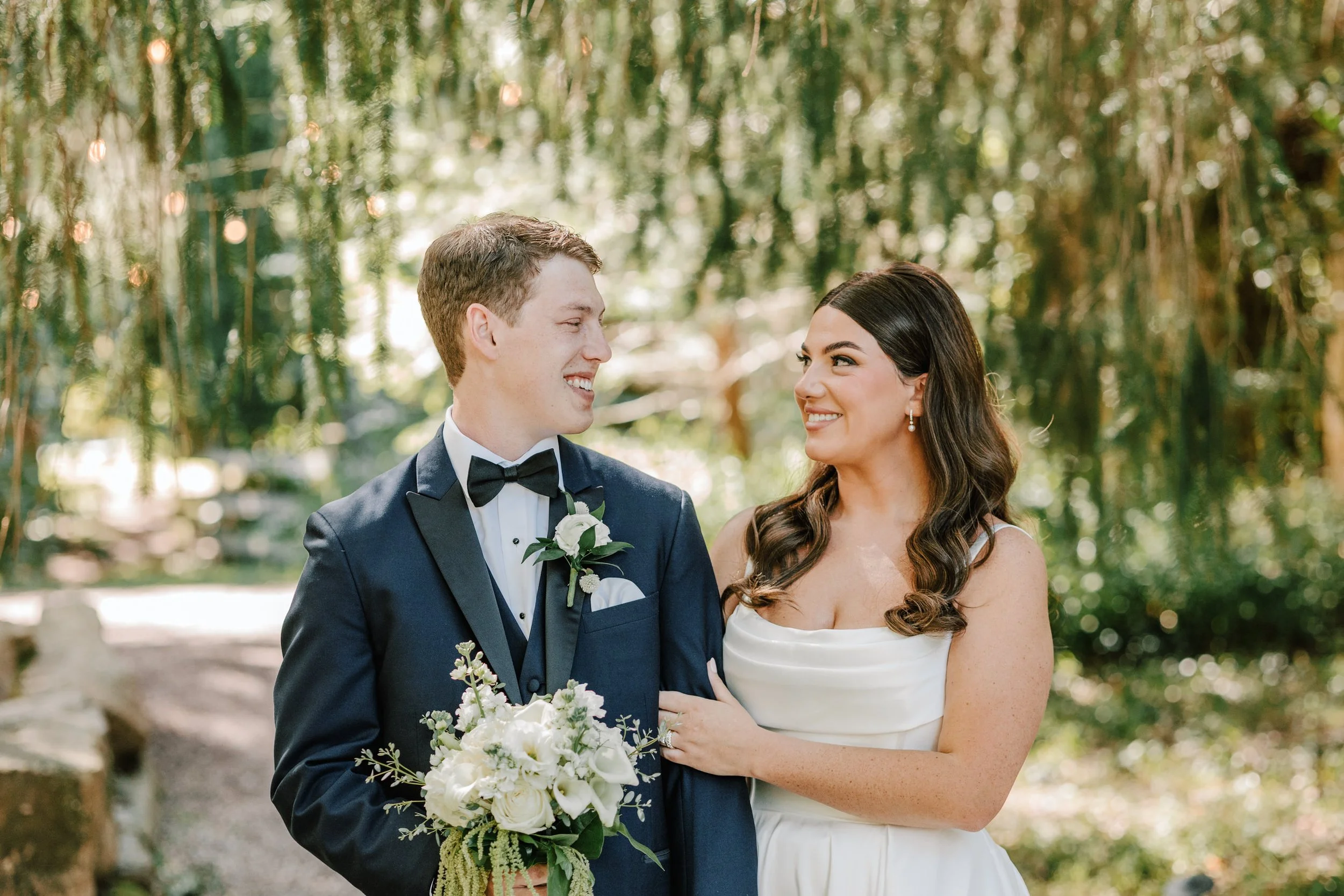 A bride and groom smiling at each other outdoors among greenery, the bride in a white strapless dress and the groom in a dark tuxedo with a bow tie, holding a white bouquet of flowers.