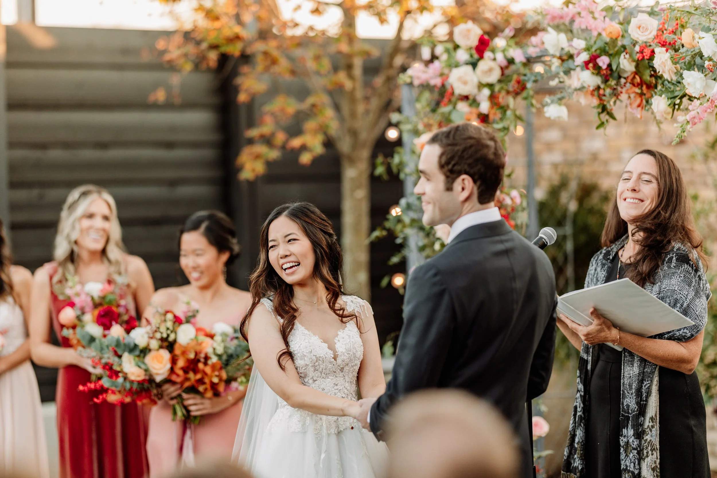 A wedding ceremony outdoors with a bride and groom holding hands, surrounded by bridesmaids, with a floral arch and autumn foliage in the background.