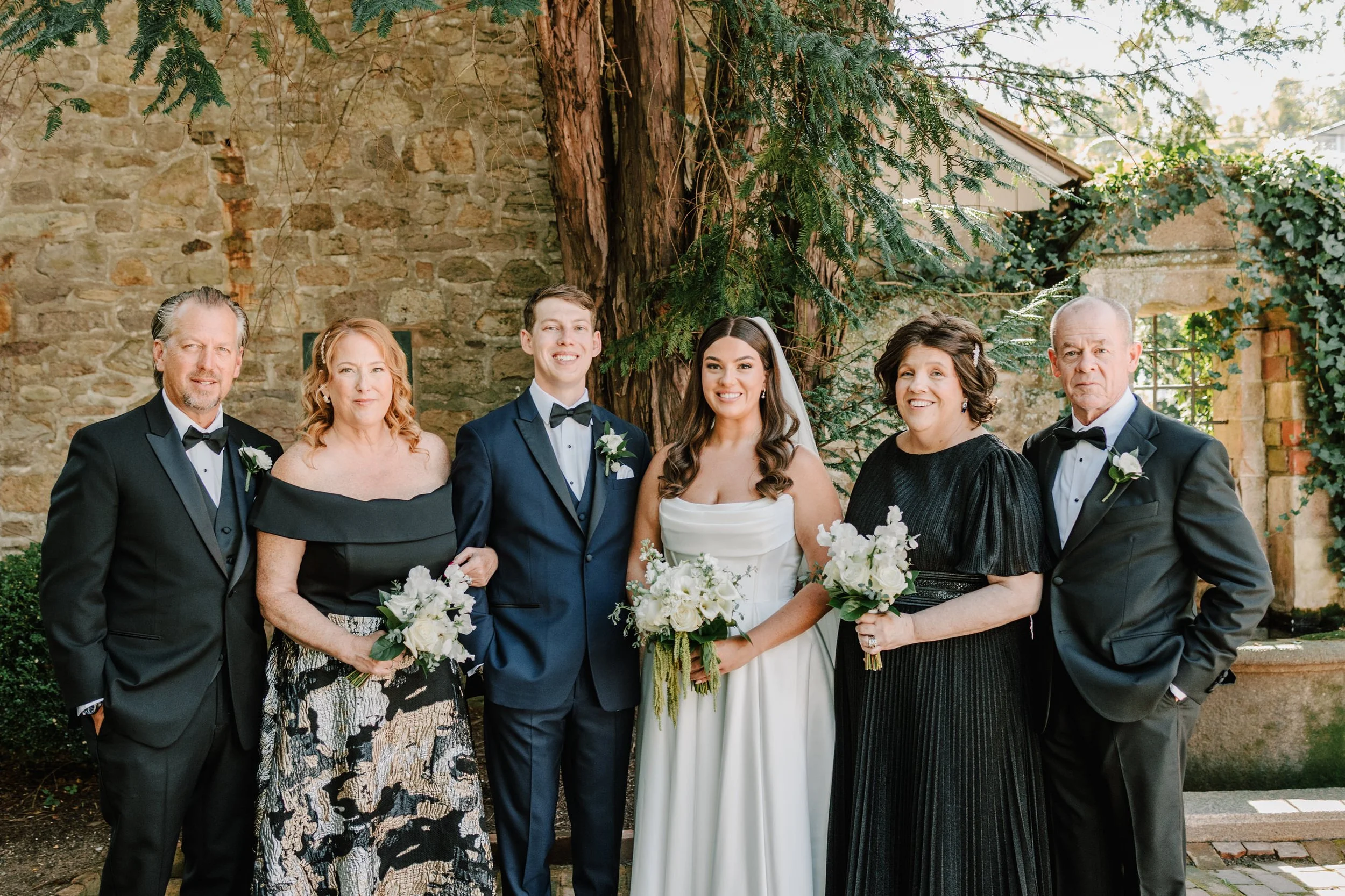 Group of six people, including bride and groom, standing outdoors against a brick wall and greenery, dressed in formal wedding attire. The bride is in a white gown holding a bouquet, and the groom in a navy suit.