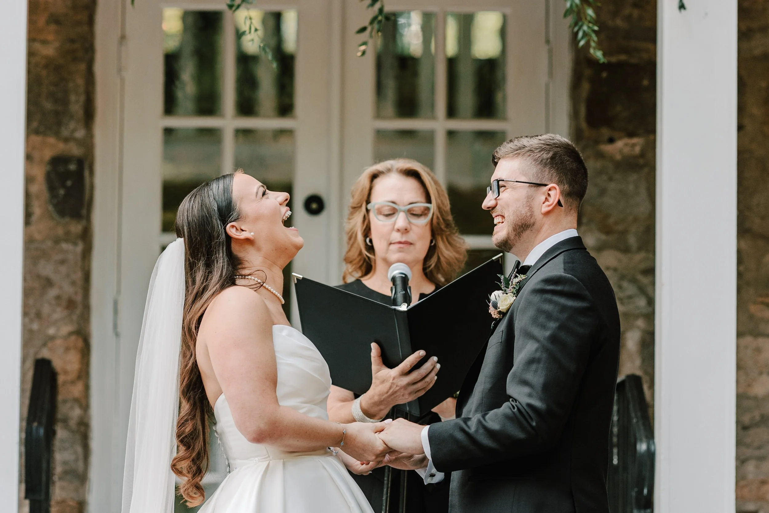 A bride and groom holding hands and smiling at each other during their wedding ceremony outdoors, with an officiant reading from a book behind them. The bride is wearing a white strapless dress and veil, while the groom is in a black tuxedo with glas