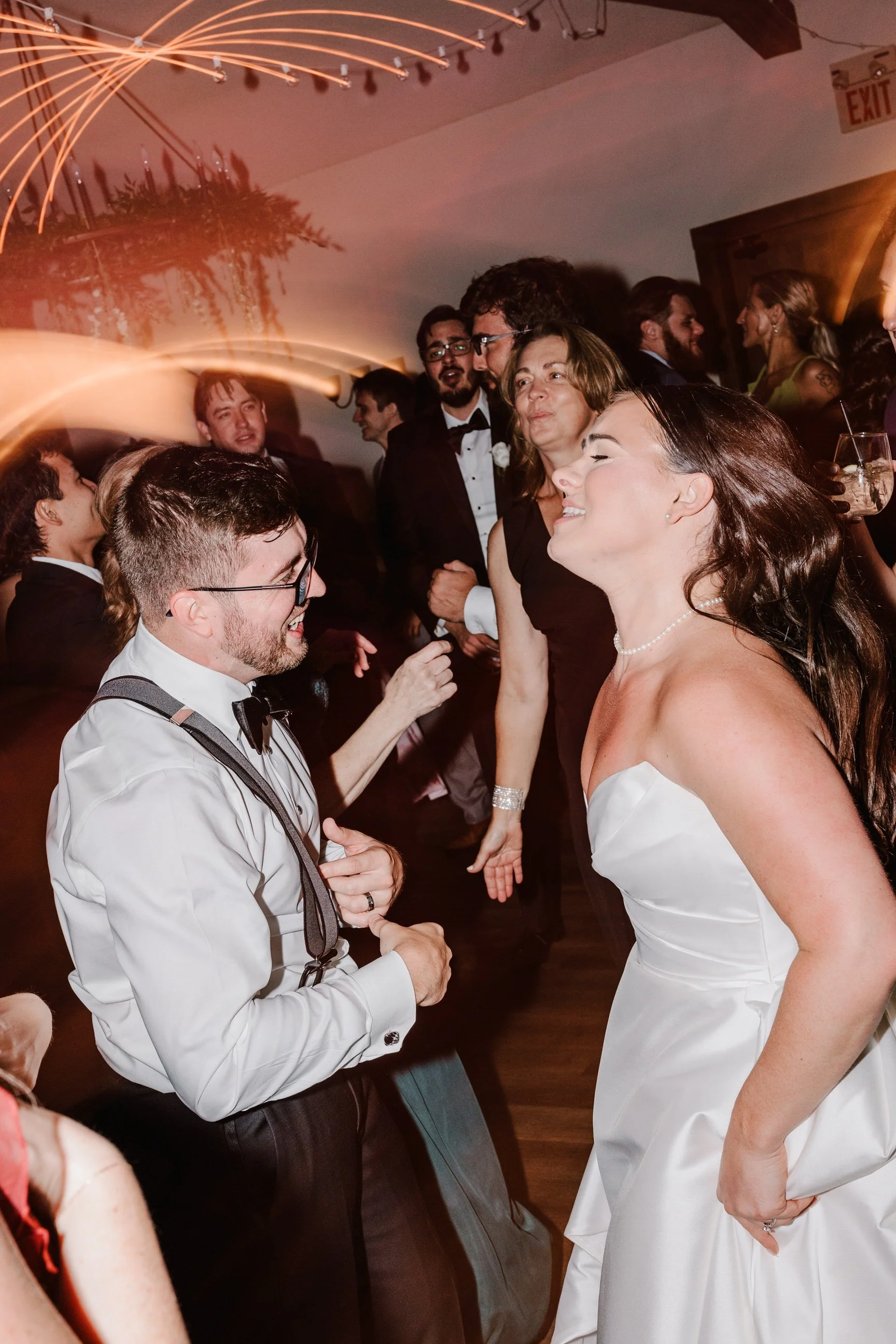 A bride and a groom dancing and laughing at their wedding reception with other guests in the background.