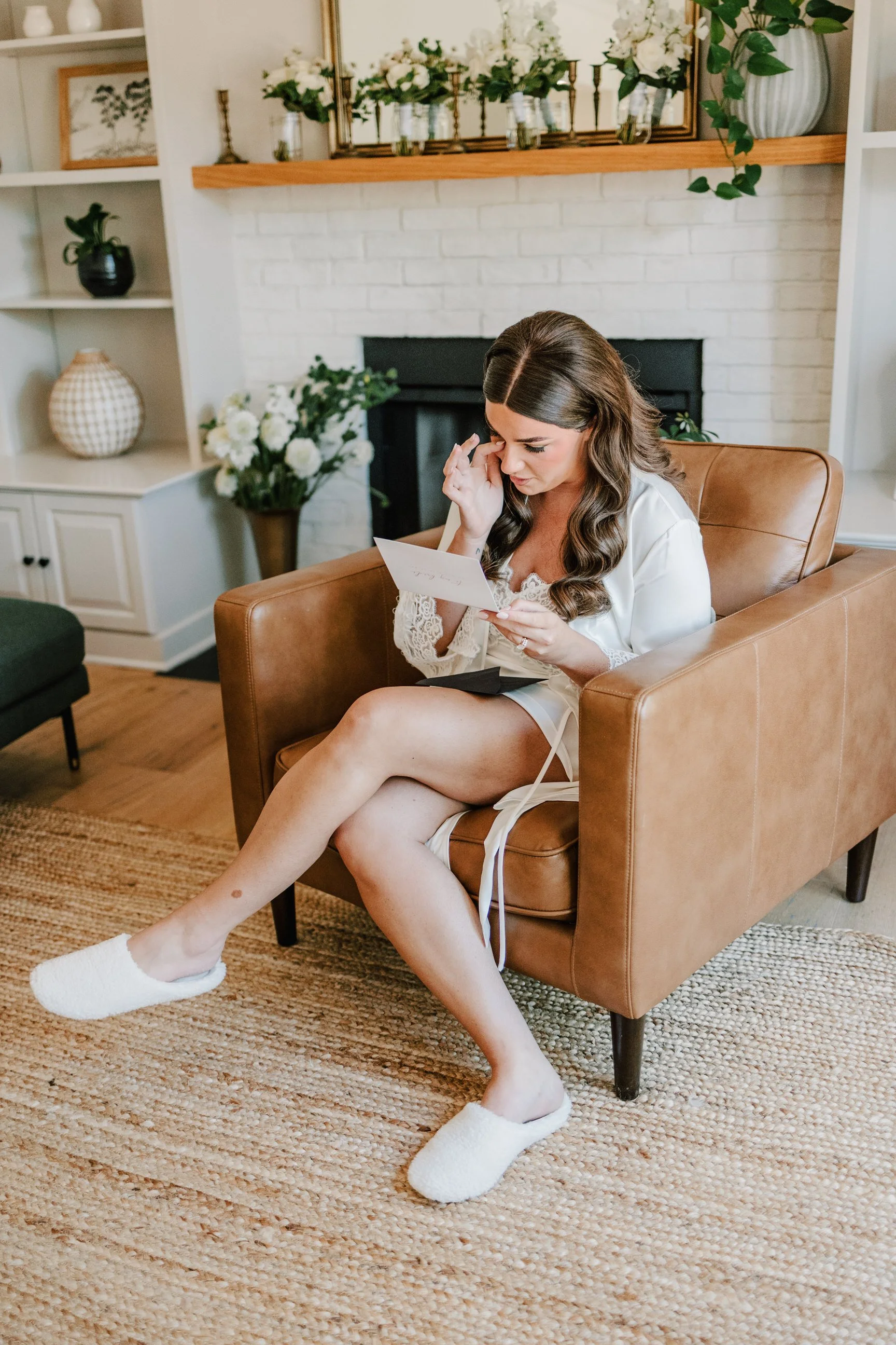 A woman sitting on a brown leather armchair in a living room, wearing a white robe and slippers, looking at a card with a surprised or emotional expression.