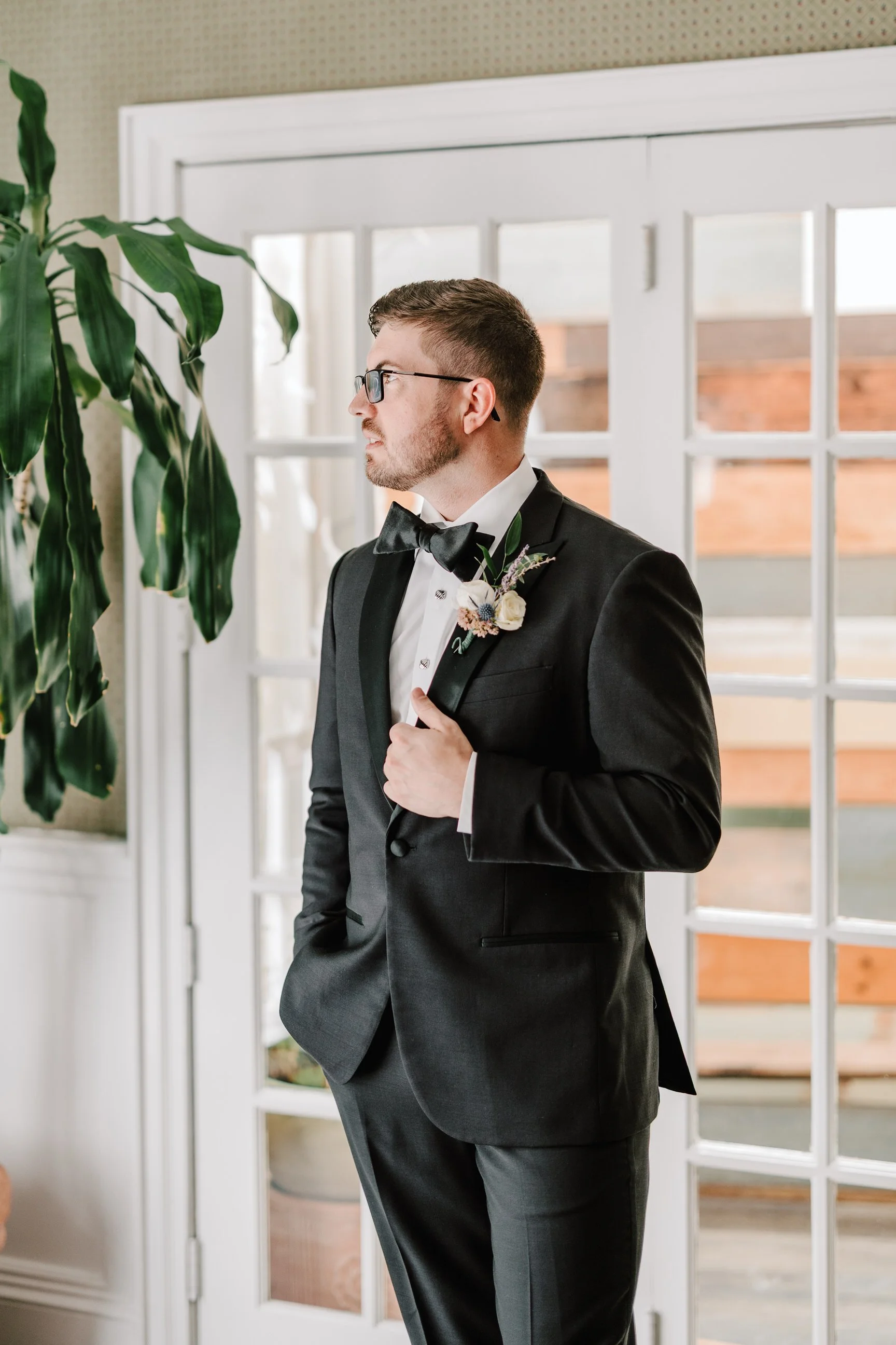 A man in a black tuxedo with a bow tie, glasses, and a boutonniere, standing indoors near a large window and a green plant.