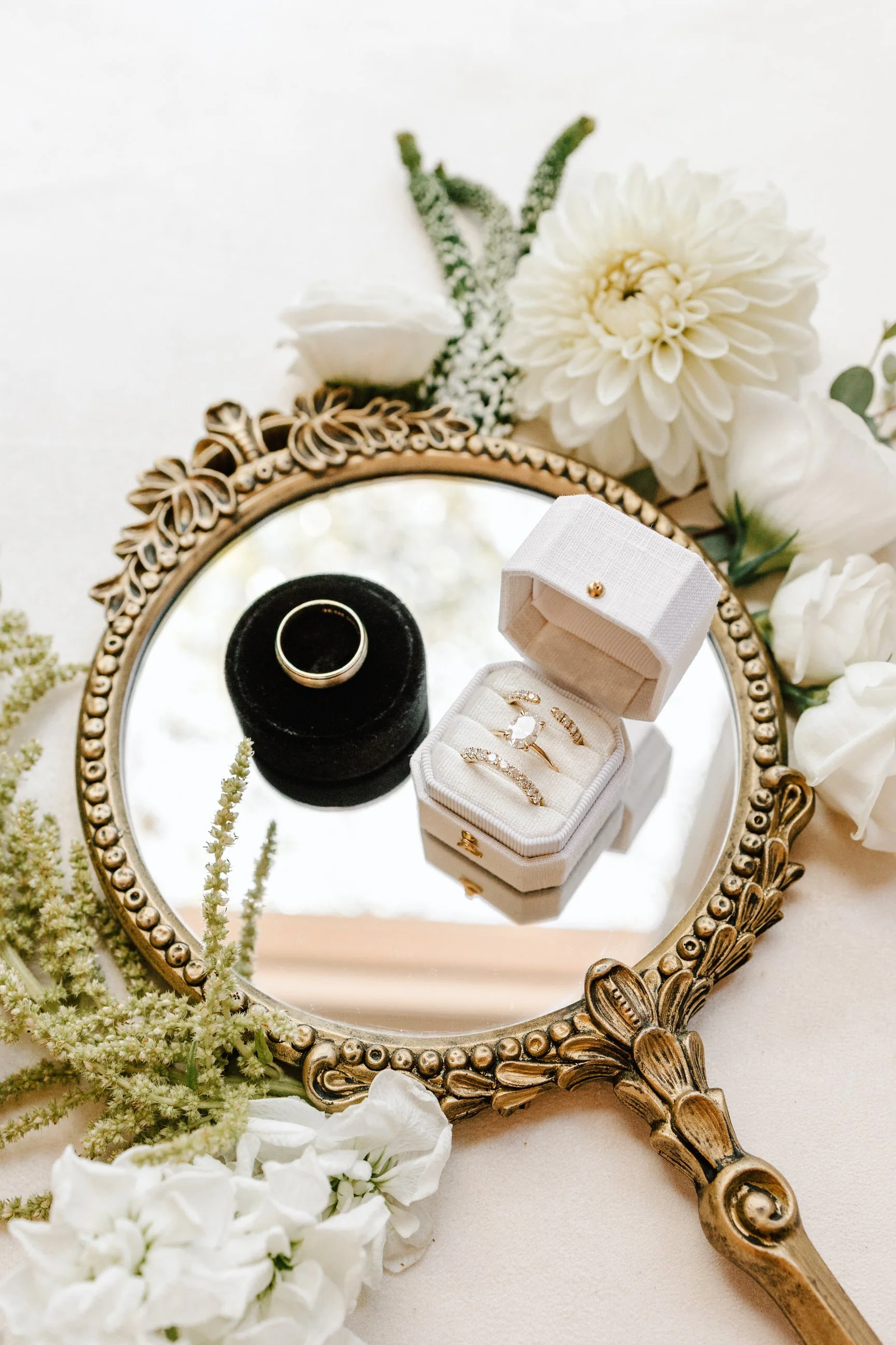 A decorative oval mirror reflects a jewelry box containing rings, a black ring holder with a silver band, surrounded by white flowers on a table, with some greenery.