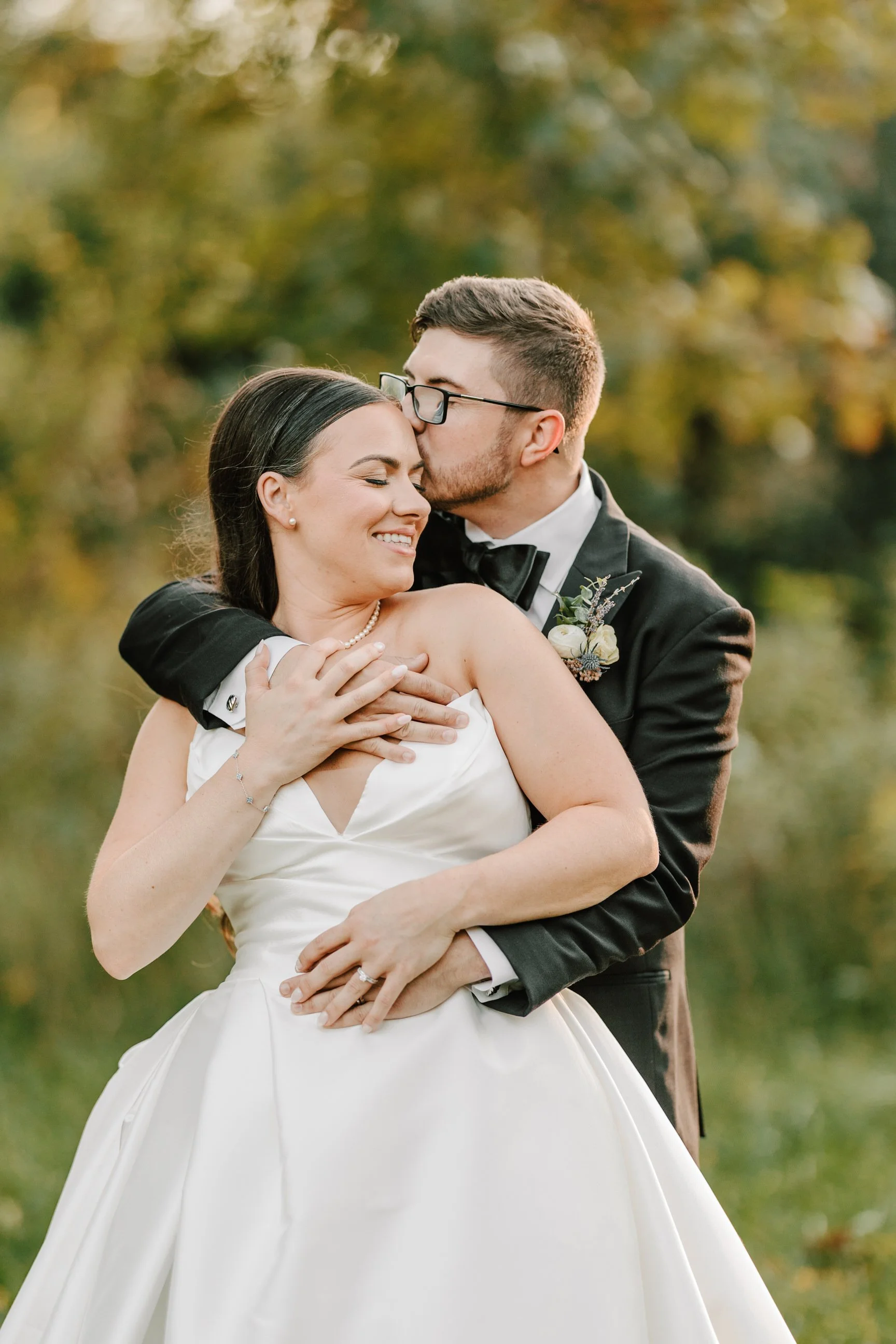 A newlywed couple embracing outdoors, with the groom kissing the bride on her forehead, as she smiles with her eyes closed. The couple is dressed in wedding attire, with a blurred natural background of trees and greenery.