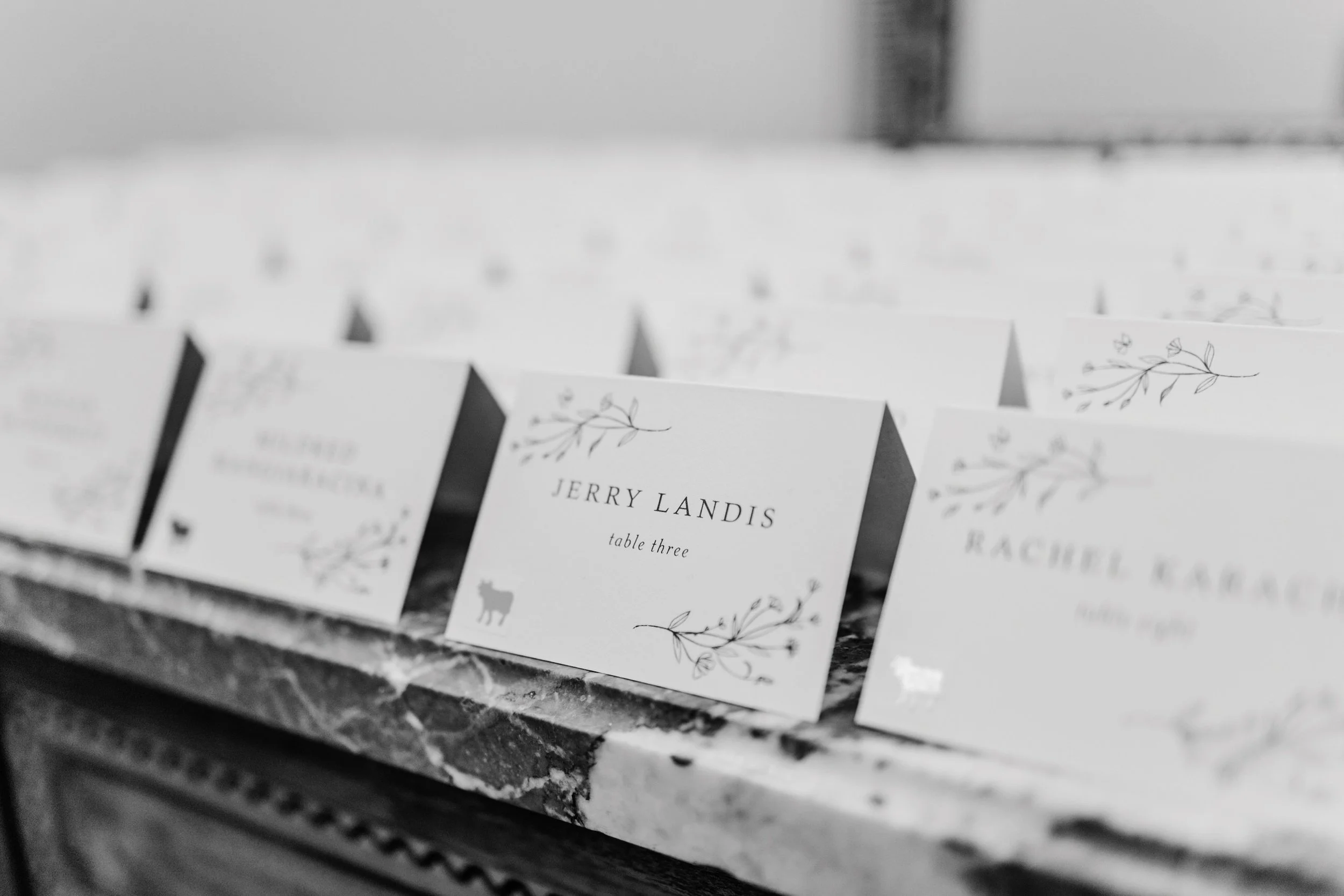 Wedding table assignment cards on a marble table, with one card reading 'Jerry Landis, table three' and decorated with a floral design and a small reindeer silhouette.