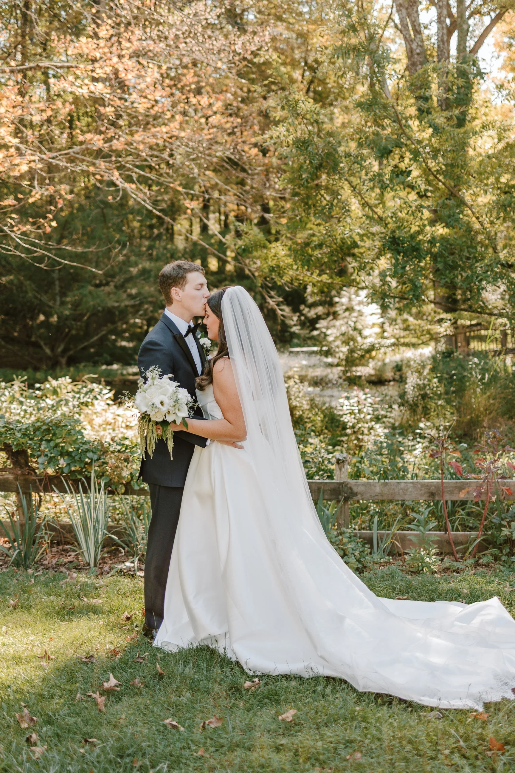 A bride and groom in wedding attire share an intimate moment outdoors, with the groom kissing the bride's forehead. The bride holds a bouquet of white flowers, and both are surrounded by trees and greenery.