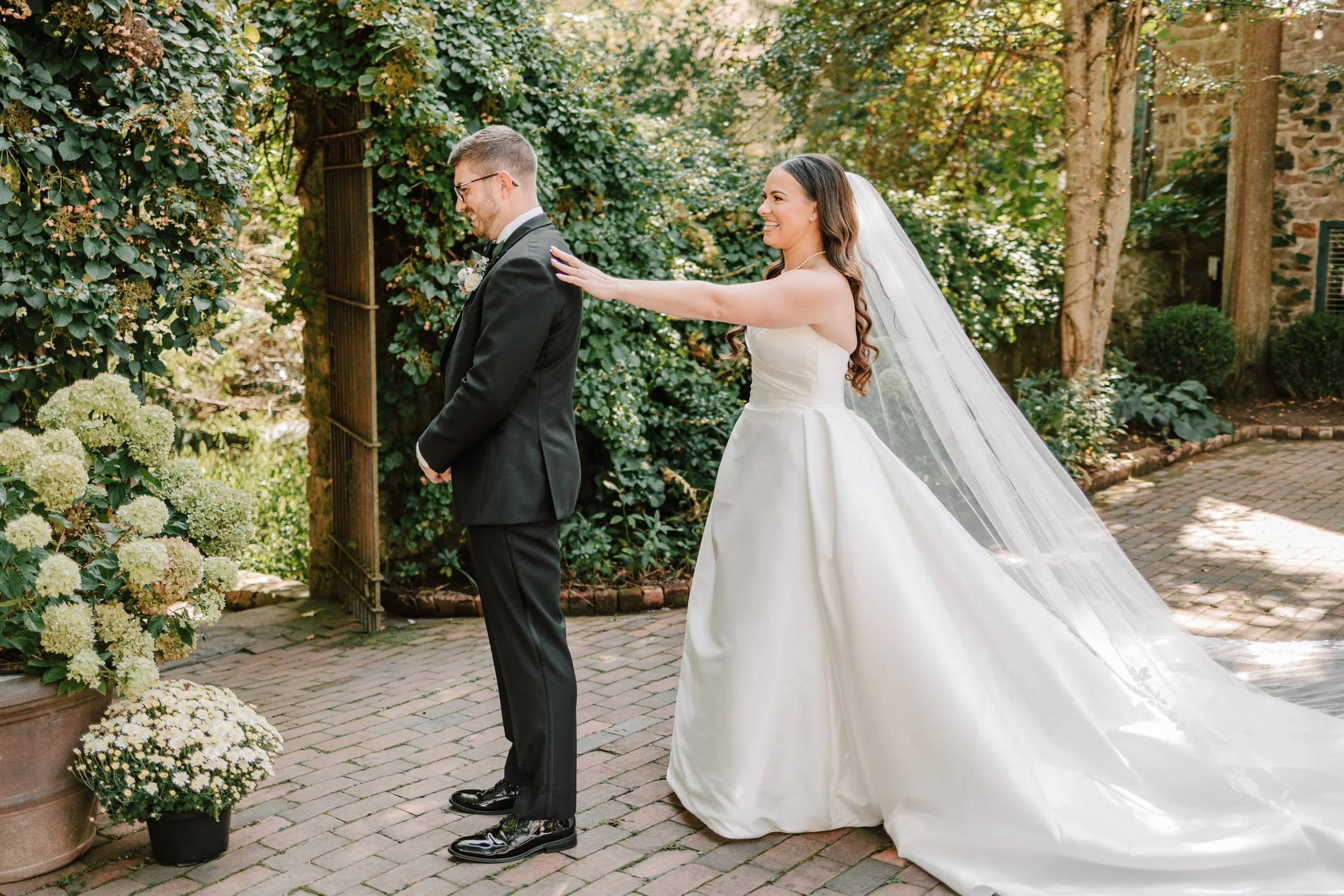 Bride in a white wedding gown and veil playfully touching groom in a black suit and tie in a garden setting with greenery and flowers.