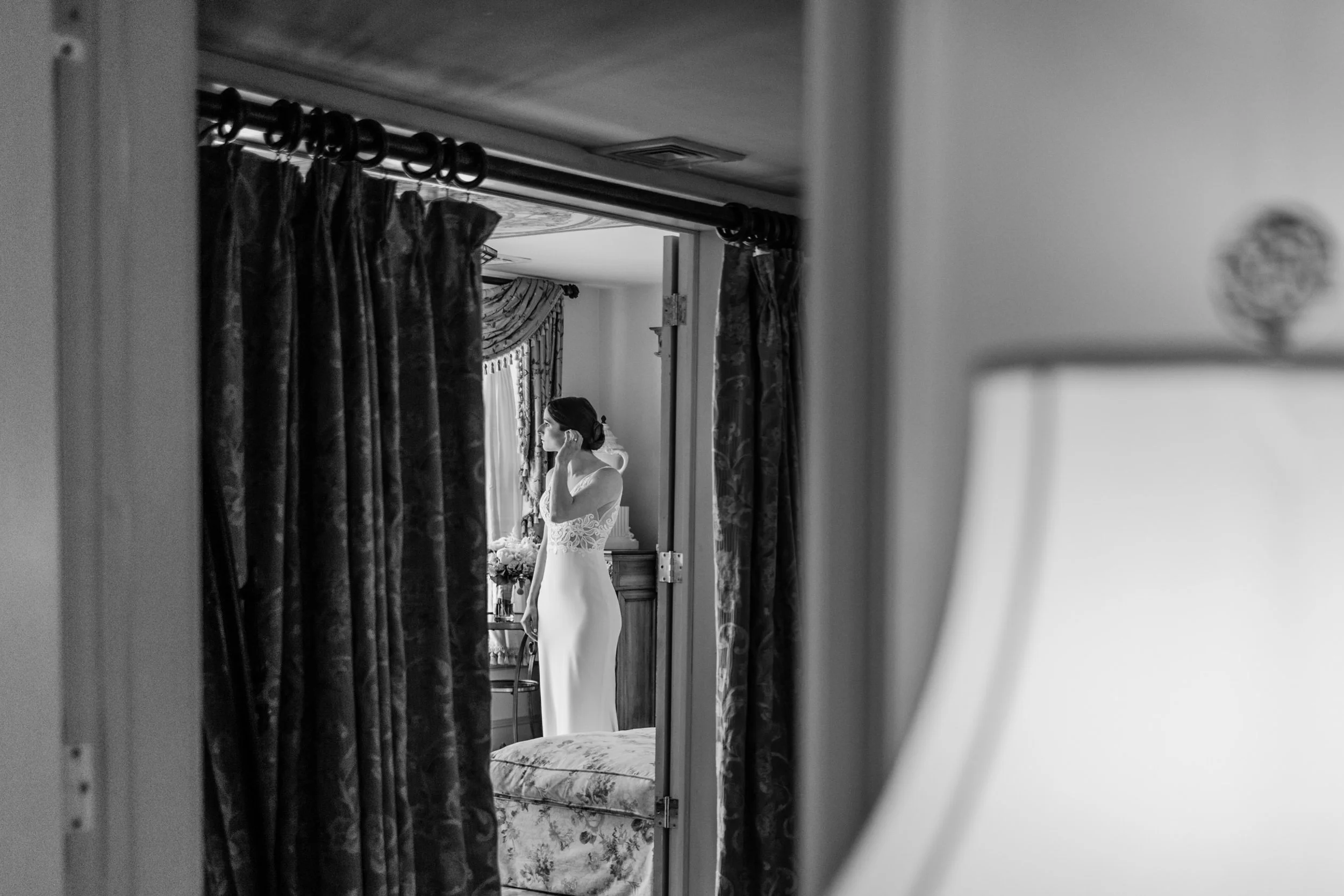 A black and white photo of a woman in a wedding dress, standing indoors by a window with curtains, seen through a doorway or mirror.