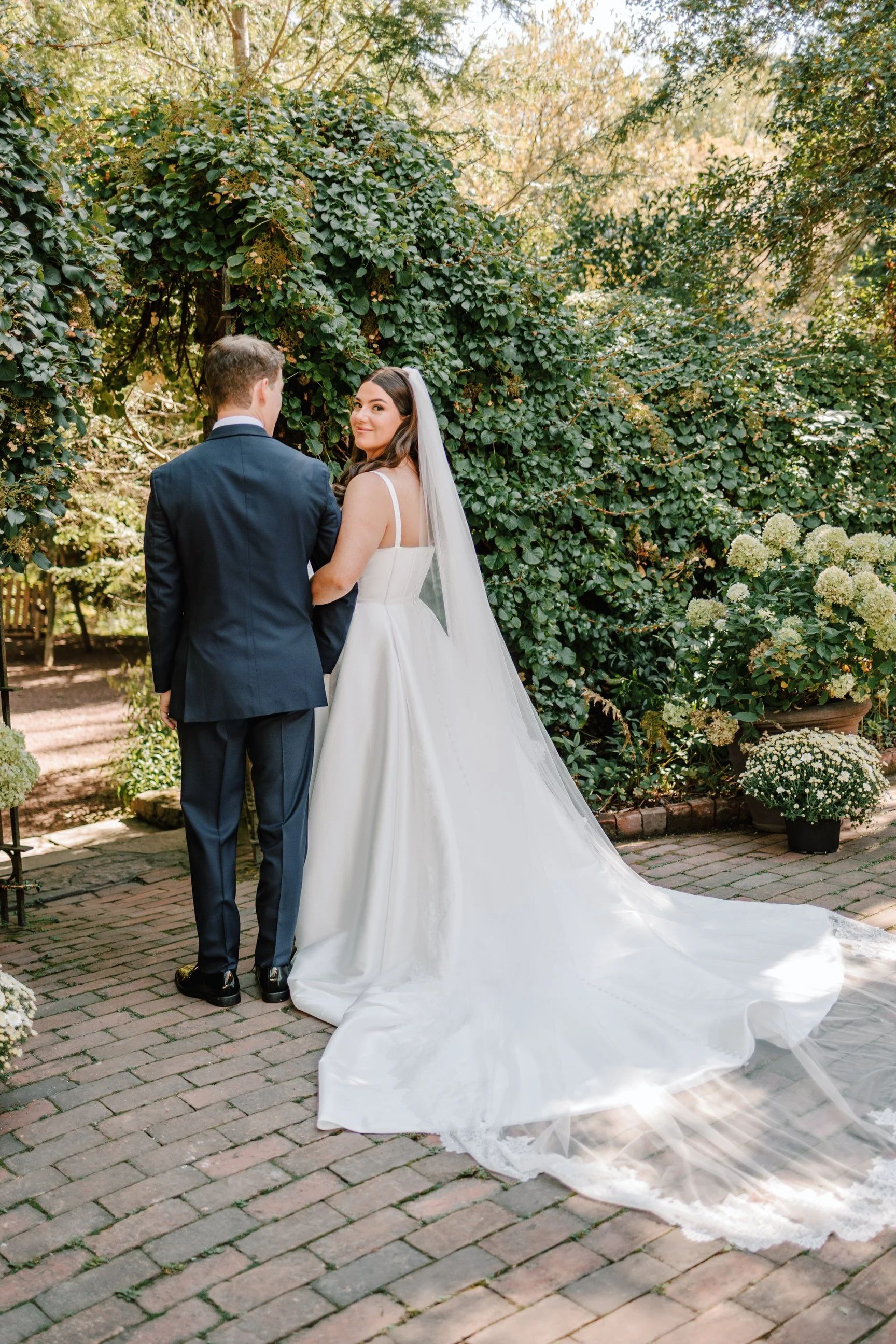 Bride and groom holding hands in a garden, with the bride looking back at the camera. The bride wears a white wedding gown with a long train and veil, while the groom wears a dark suit. Greenery and potted flowers are in the background.