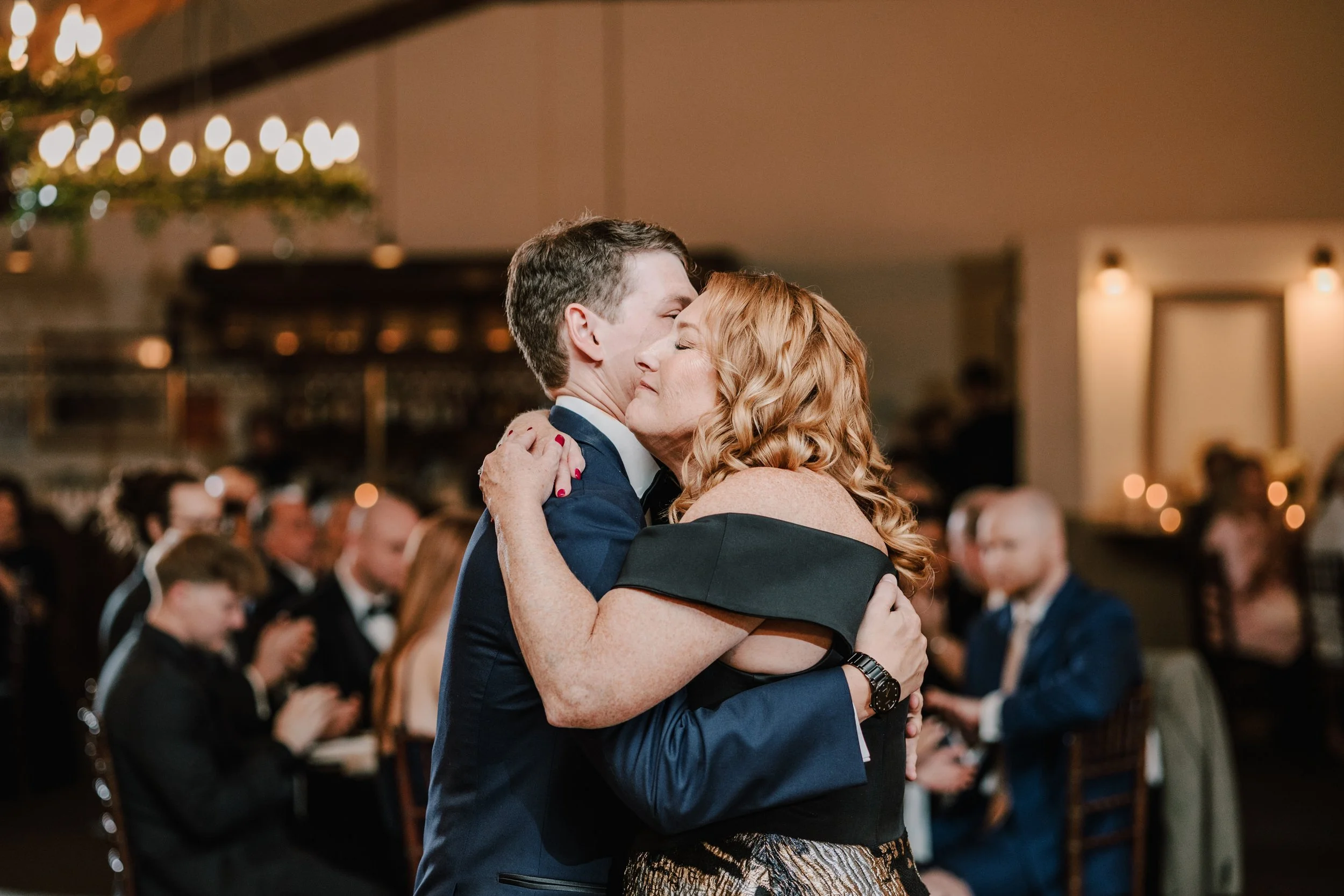 A couple is hugging at a formal event, with guests seated at tables in the background, in an elegant indoor setting with warm lighting.