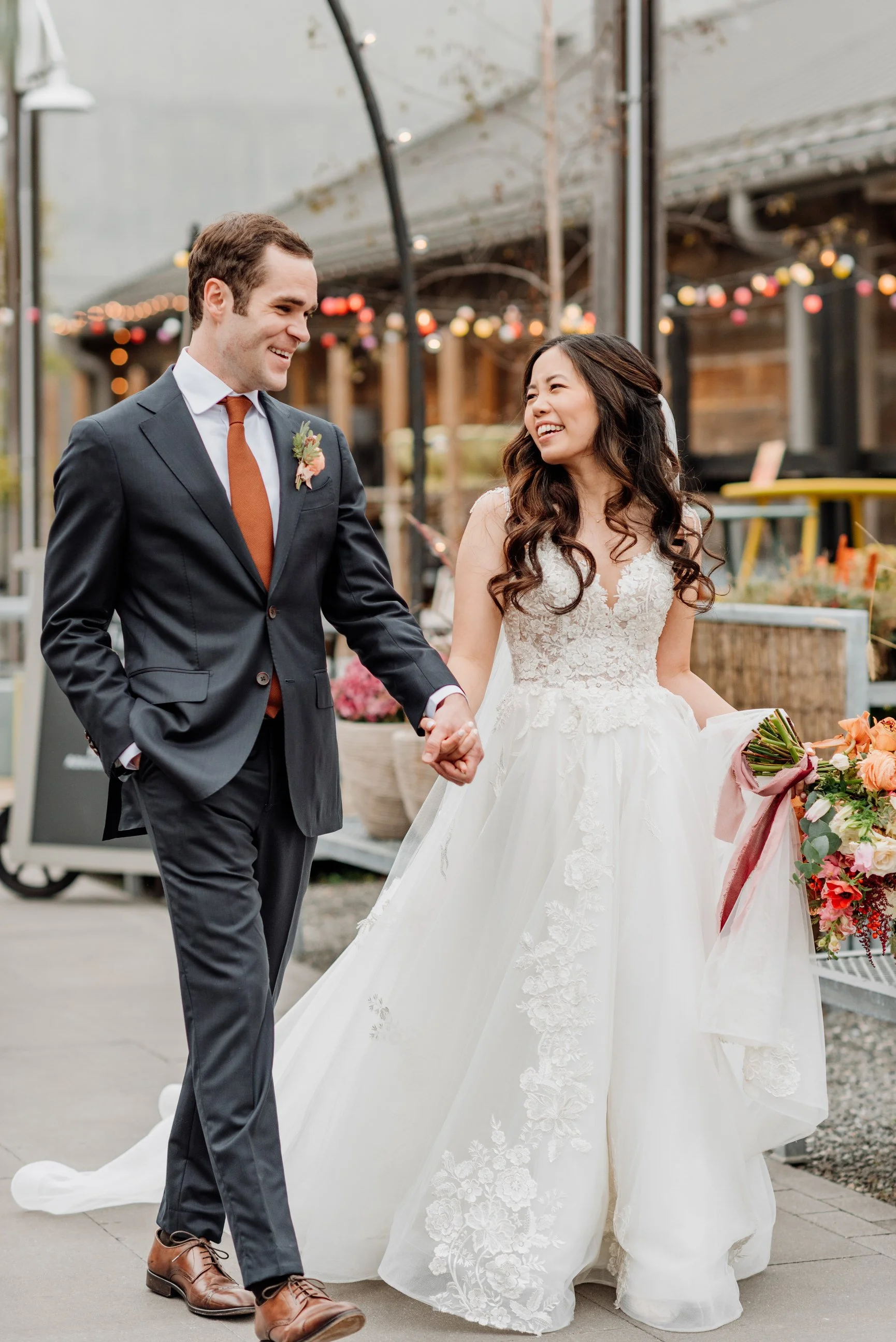 A newlywed couple holding hands and smiling at each other outdoors during their wedding reception, with string lights and floral arrangements in the background.