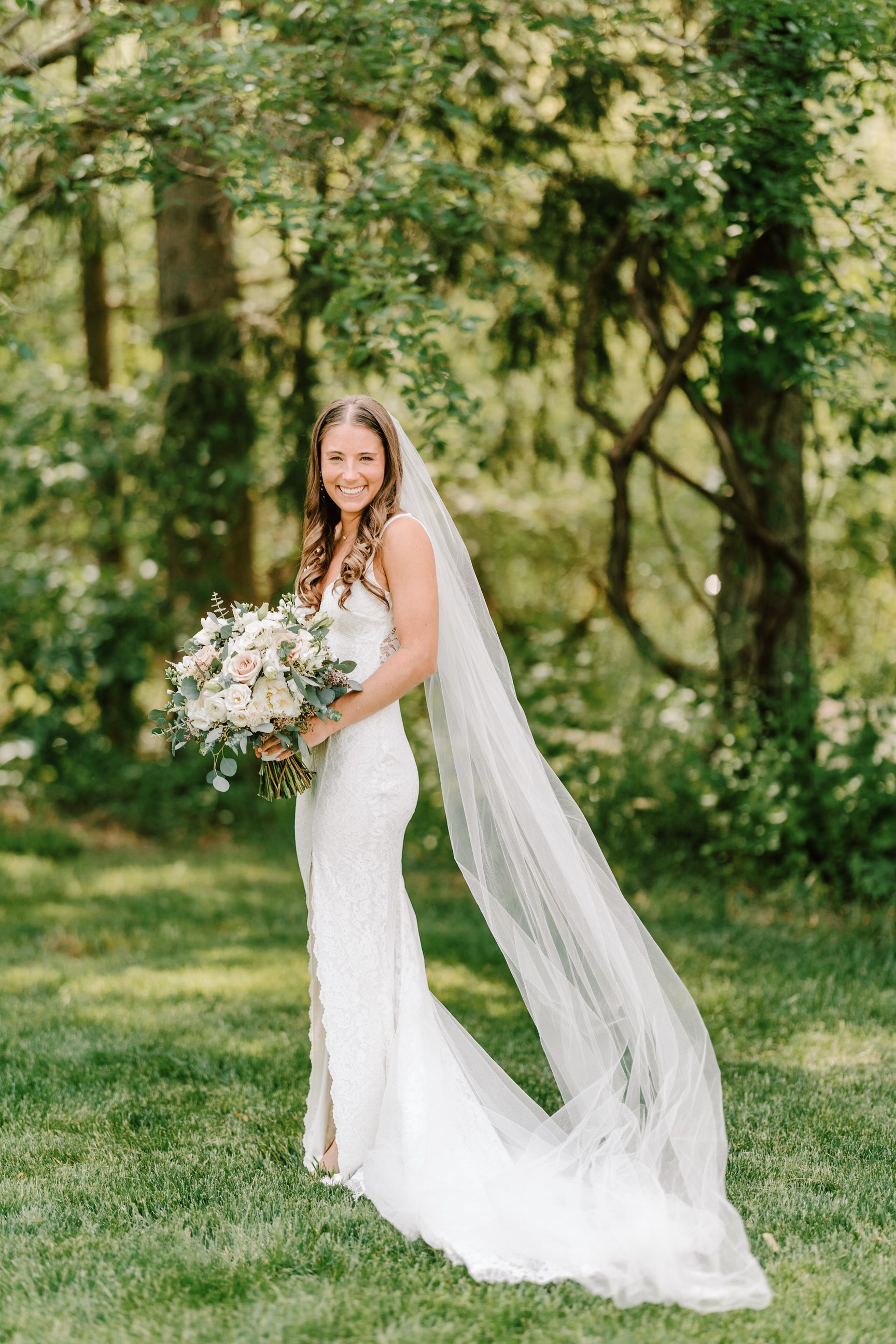 A smiling bride in a white lace wedding gown with a long veil holding a bouquet of white and blush pink flowers standing outdoors on green grass surrounded by trees.
