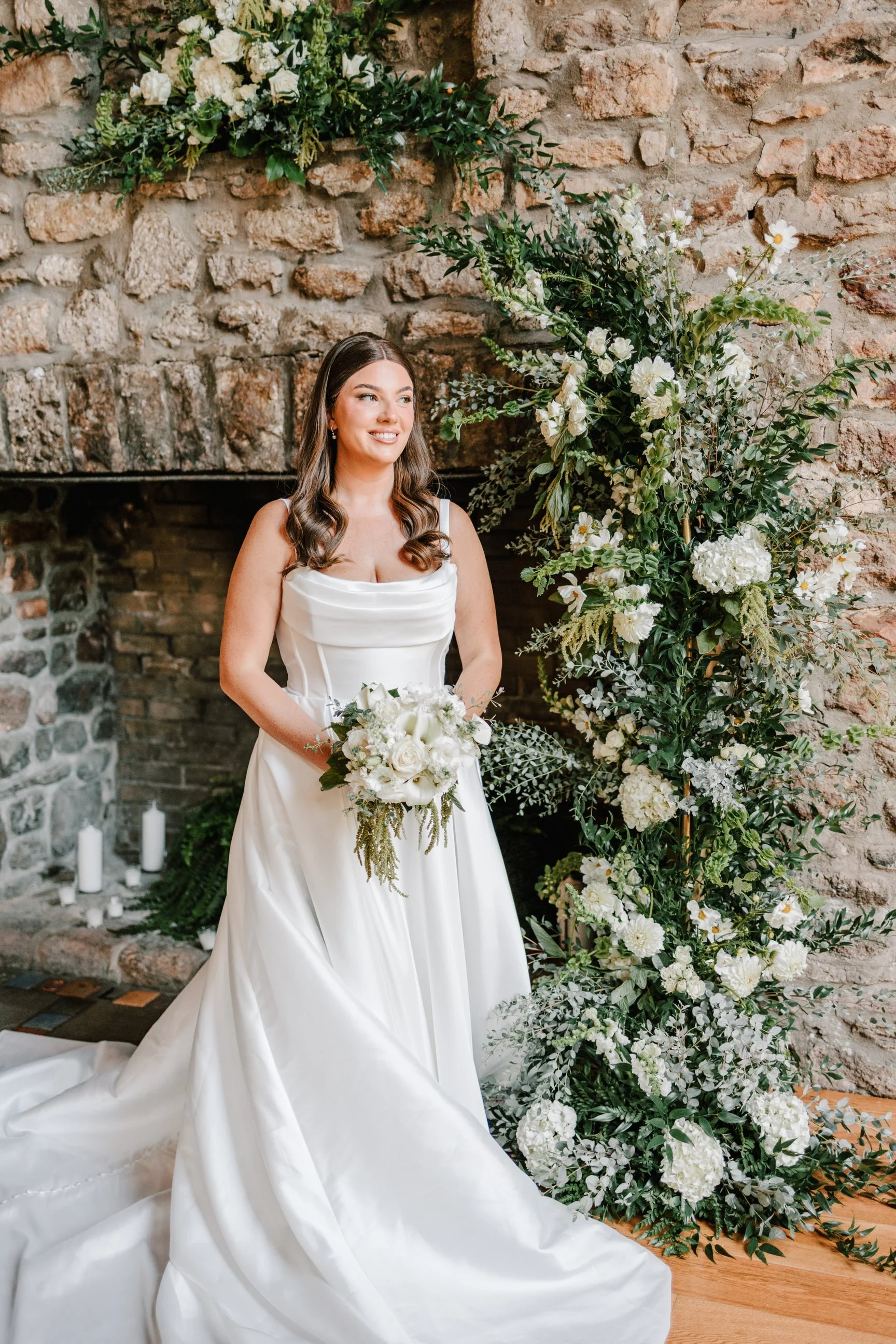 A bride in a white wedding dress holding a bouquet of white flowers, standing next to a floral arrangement with white flowers and greenery against a brick wall.