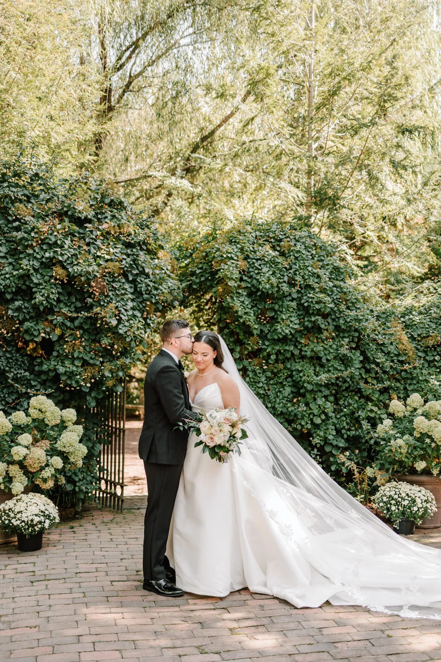 A bride and groom in wedding attire standing beneath a large green archway, with the groom kissing the bride's forehead. The bride holds a bouquet of pink and white flowers, and both are smiling peacefully.