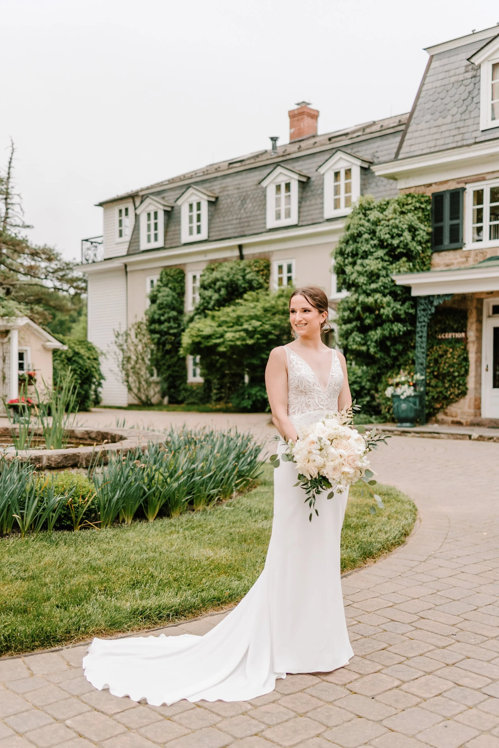 A bride in a white wedding dress holding a bouquet of white flowers stands outdoors on a paved path in front of a large house with lush green trees and shrubs.