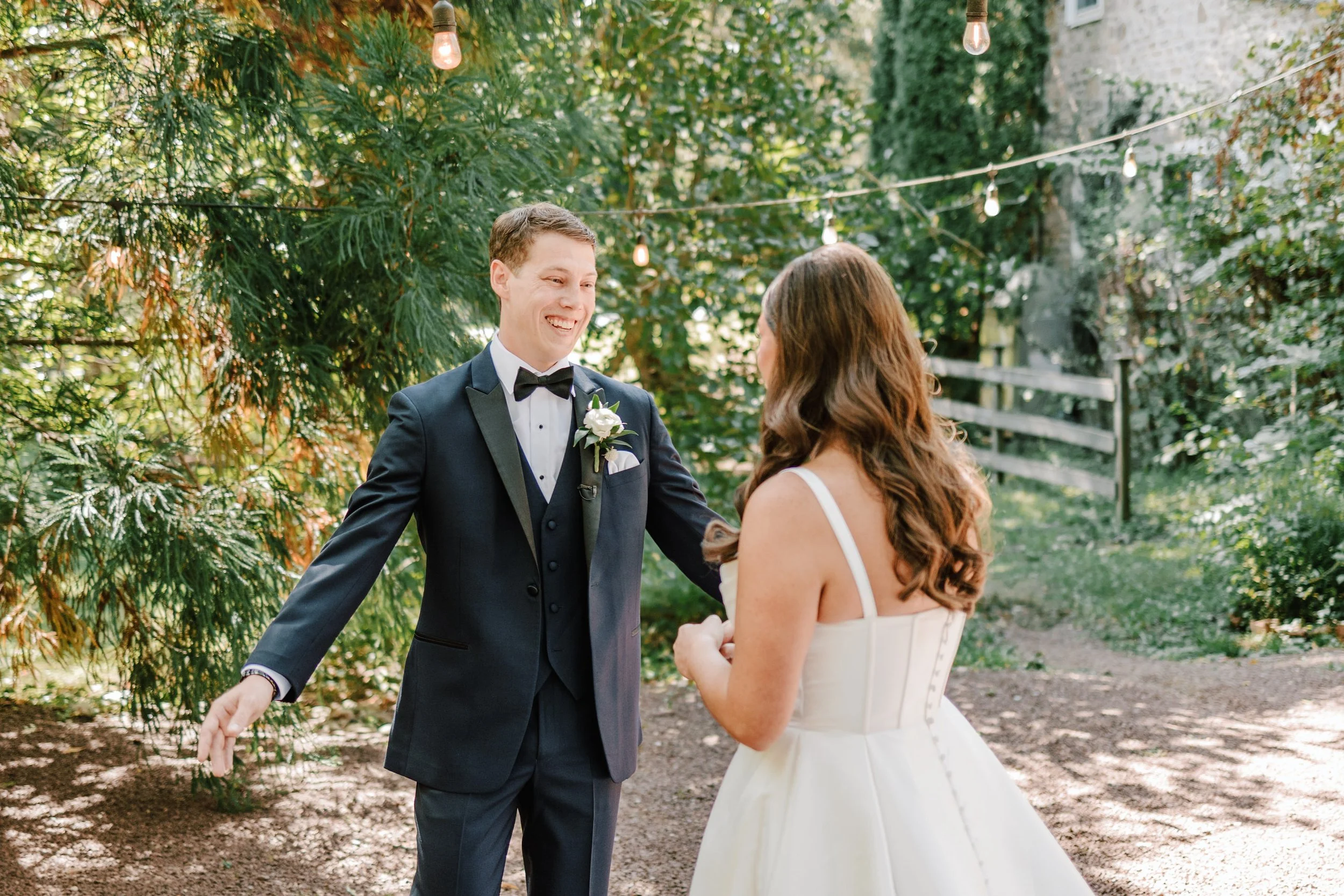 A groom in a black tuxedo and a bride in a white wedding dress are standing outdoors, smiling at each other, with string lights and lush greenery in the background.