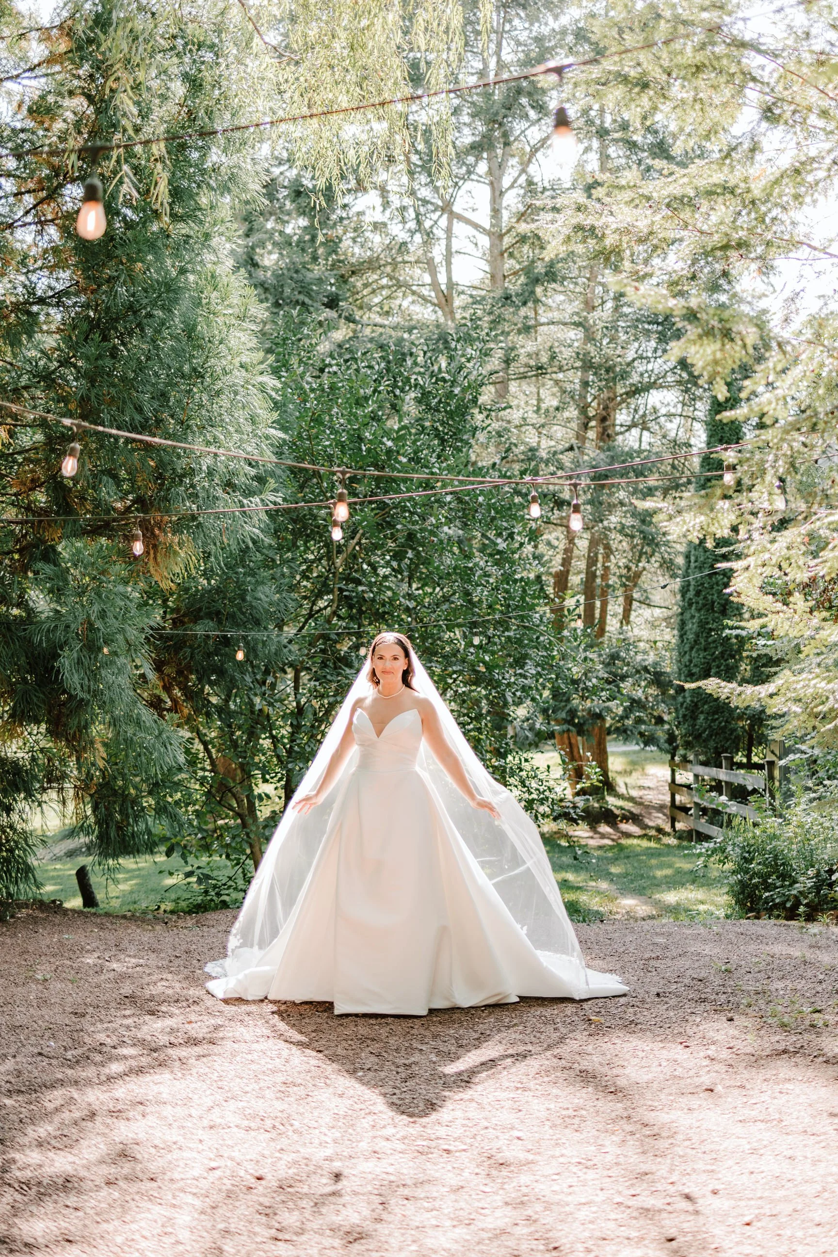 Bride in a white wedding gown standing outdoors under string lights, surrounded by trees and greenery.
