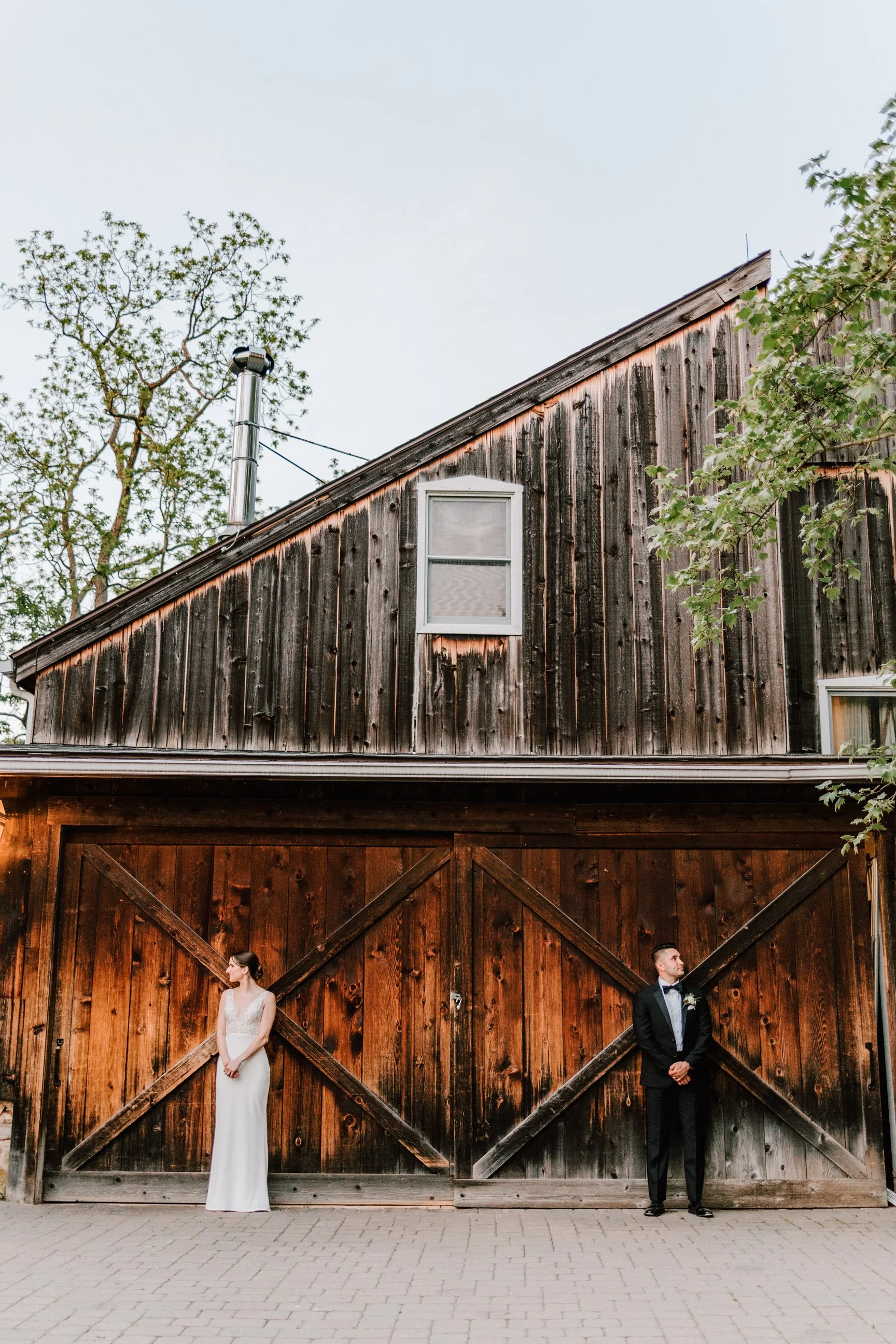 A bride in a white wedding dress and a groom in a black tuxedo stand apart in front of a large wooden barn.