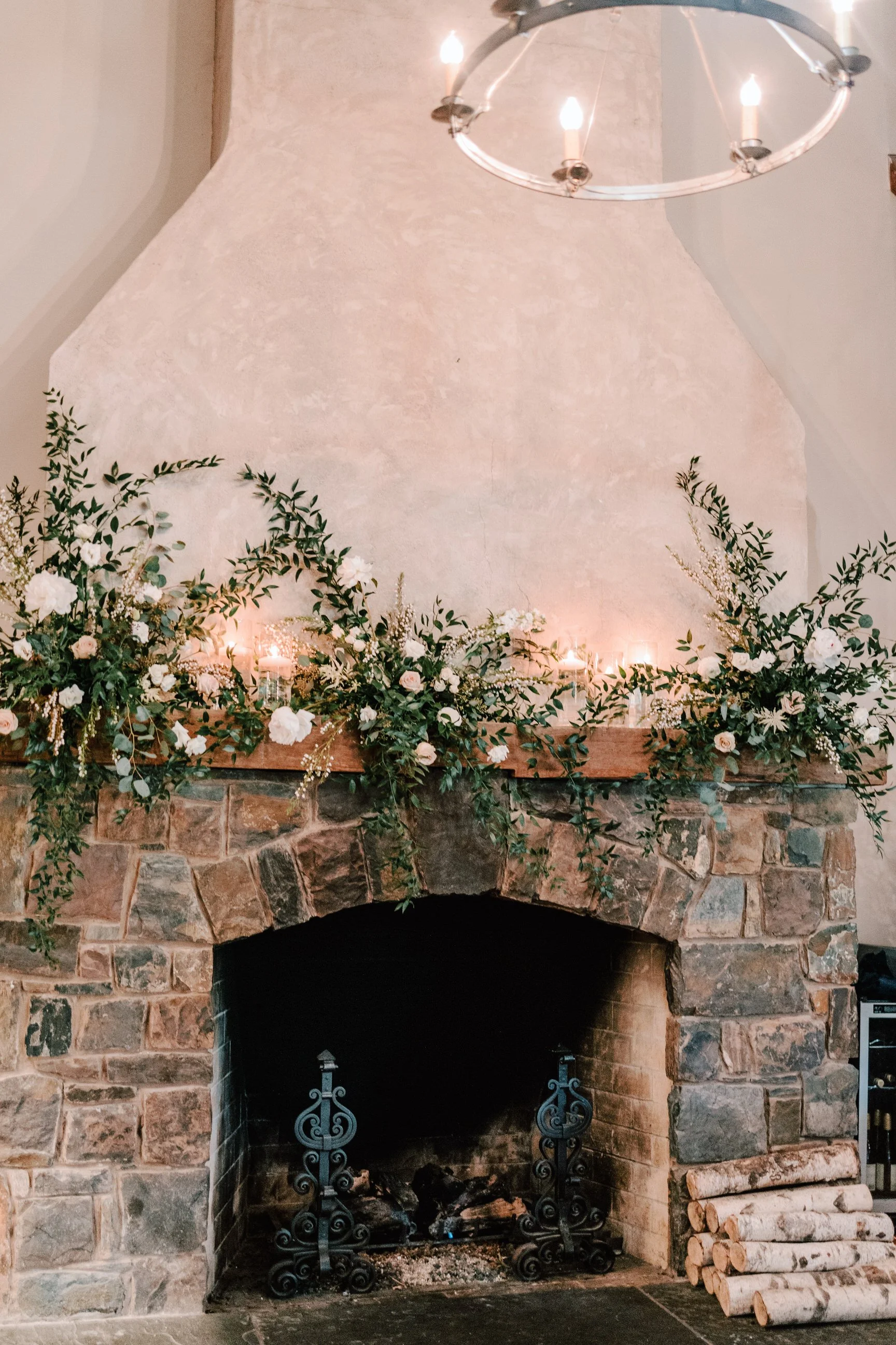 Decorative stone fireplace with a floral and greenery mantel arrangement, candles, and a wooden log pile on the right, under a circular chandelier.