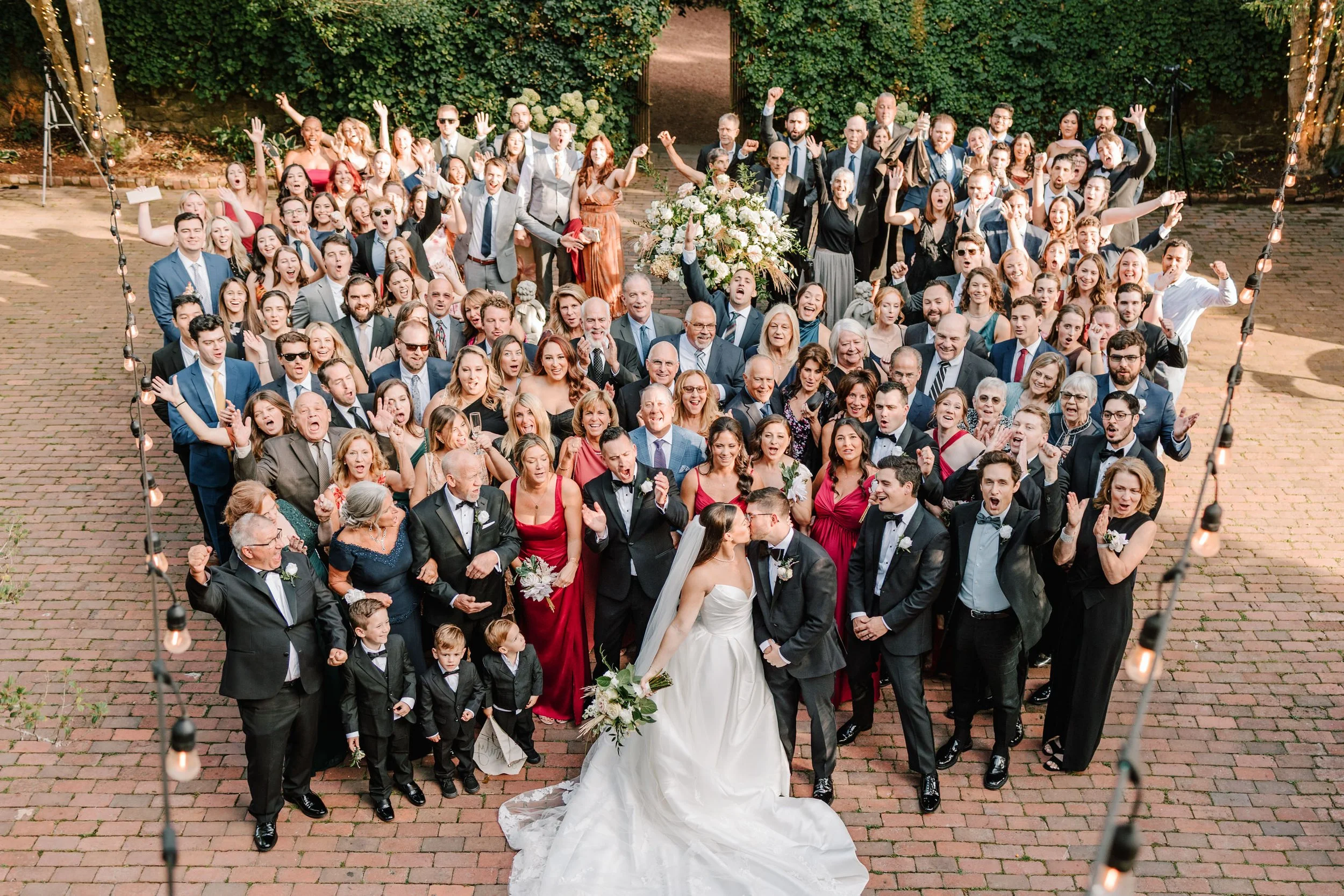 Large group of wedding guests gathered outdoors on a brick courtyard, celebrating a wedding, with a bride and groom kissing in the front center surrounded by friends and family.