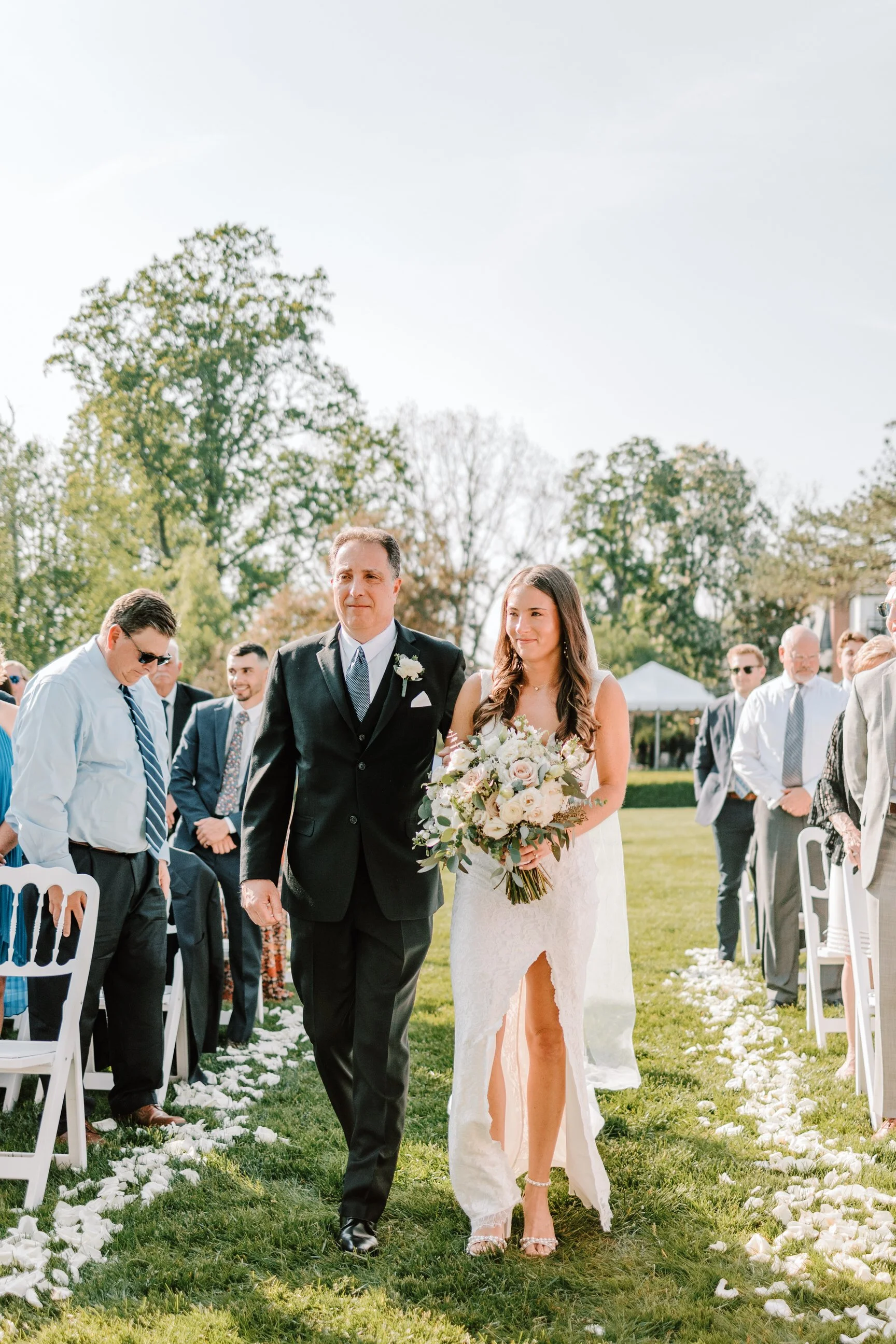 A bride walking down the aisle with her father at an outdoor wedding ceremony. The bride is holding a bouquet of flowers and wearing a lace wedding dress with a slit, while the father is dressed in a black suit with a white shirt and tie. Guests are 