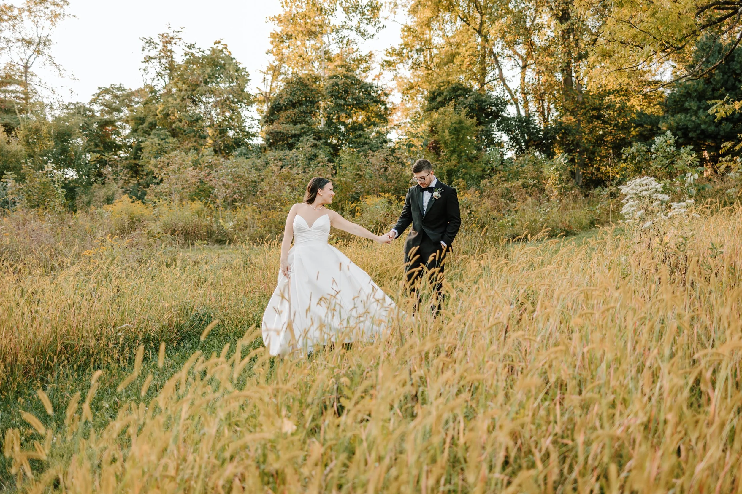 A bride in a white dress and a groom in a black tuxedo holding hands in a field of tall, golden grass with trees in the background during autumn.